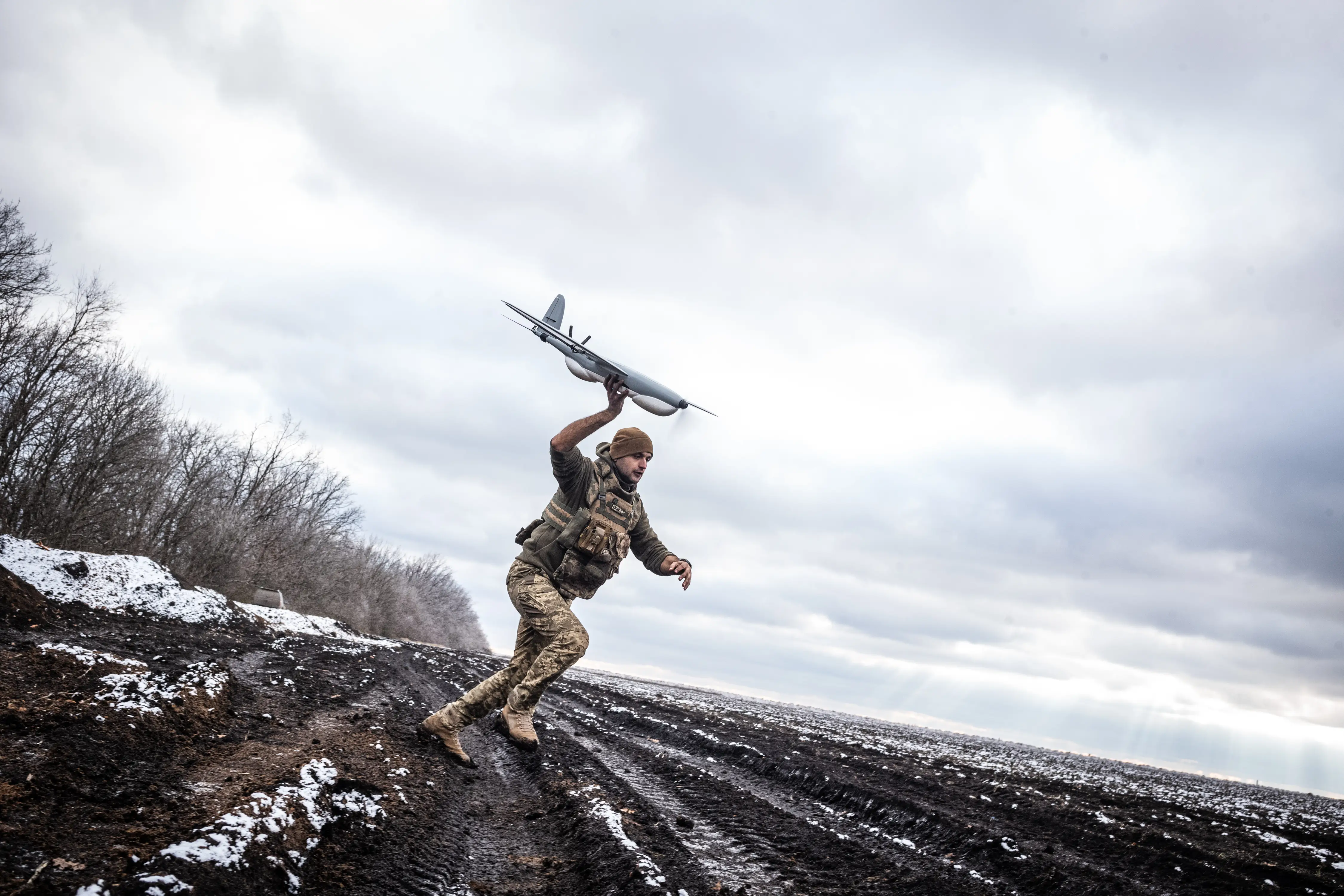 A man in camouflage gear runs on a muddy and snowy field while holding a grey drone in the sky, under a grey sky and with a few trees behind him