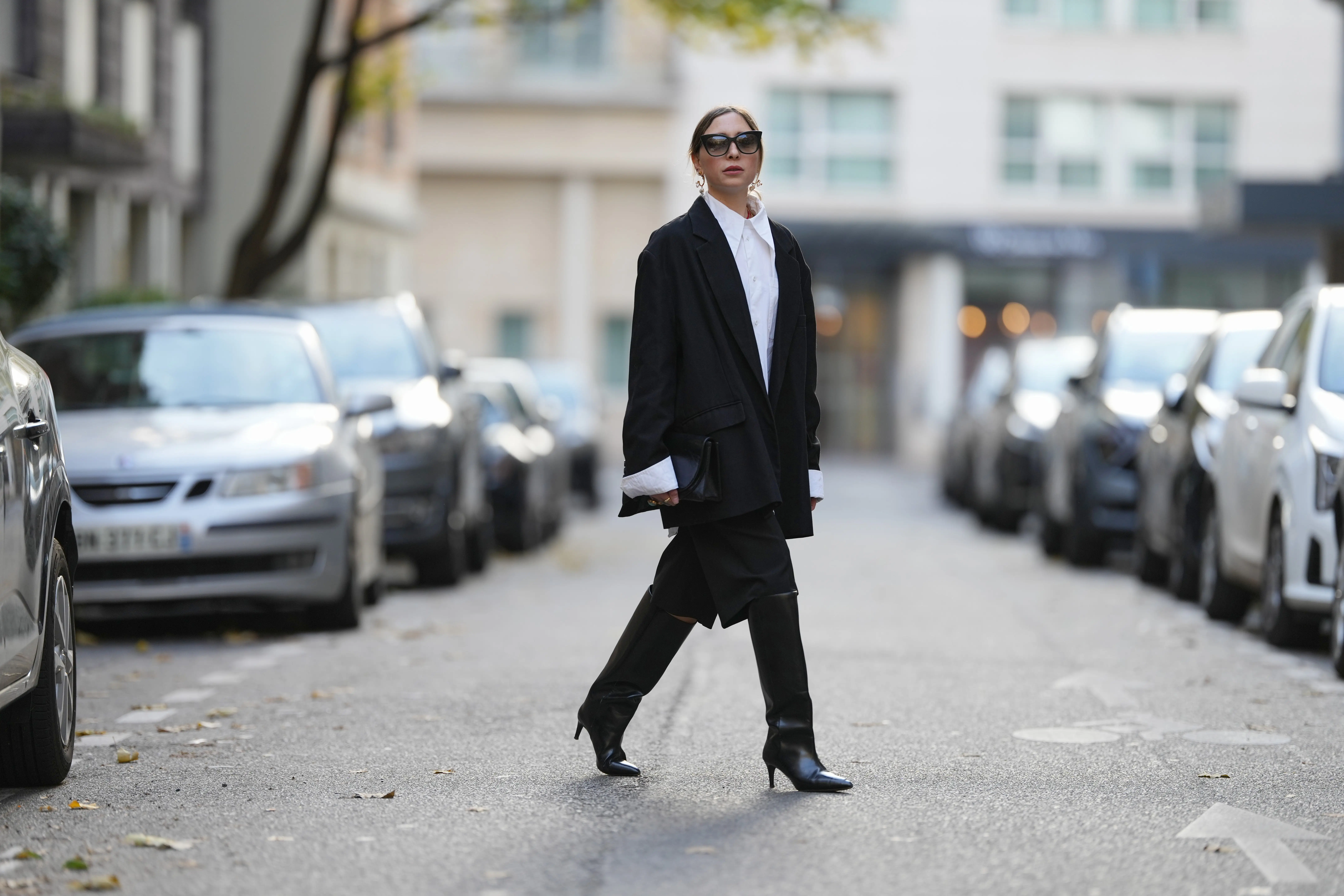 Emy Venturini walks across the road between parked cars wearing black leather heeled boots by Toteme.