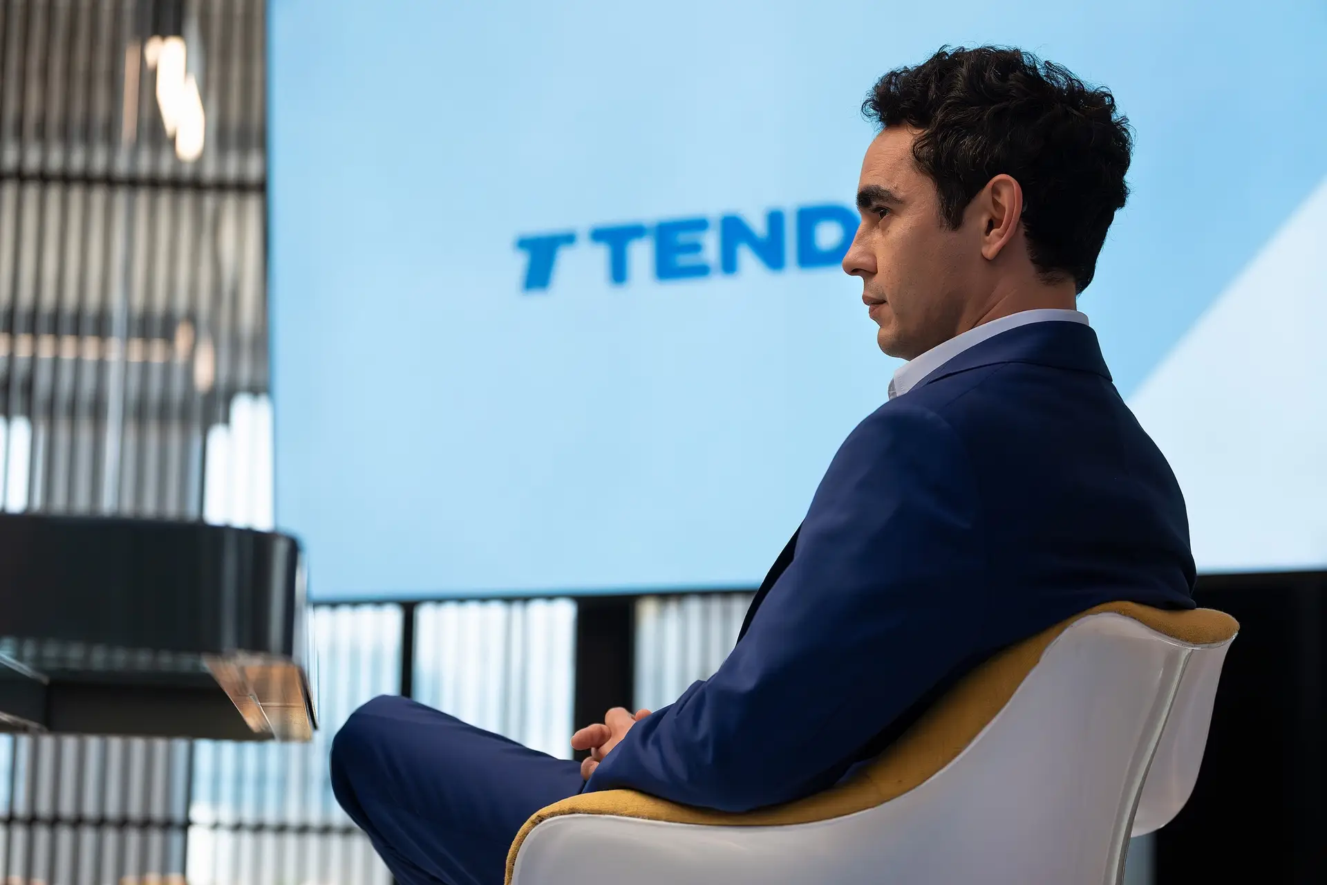 A man sits in side profile in a suit in front of a screen for a company called Tender.