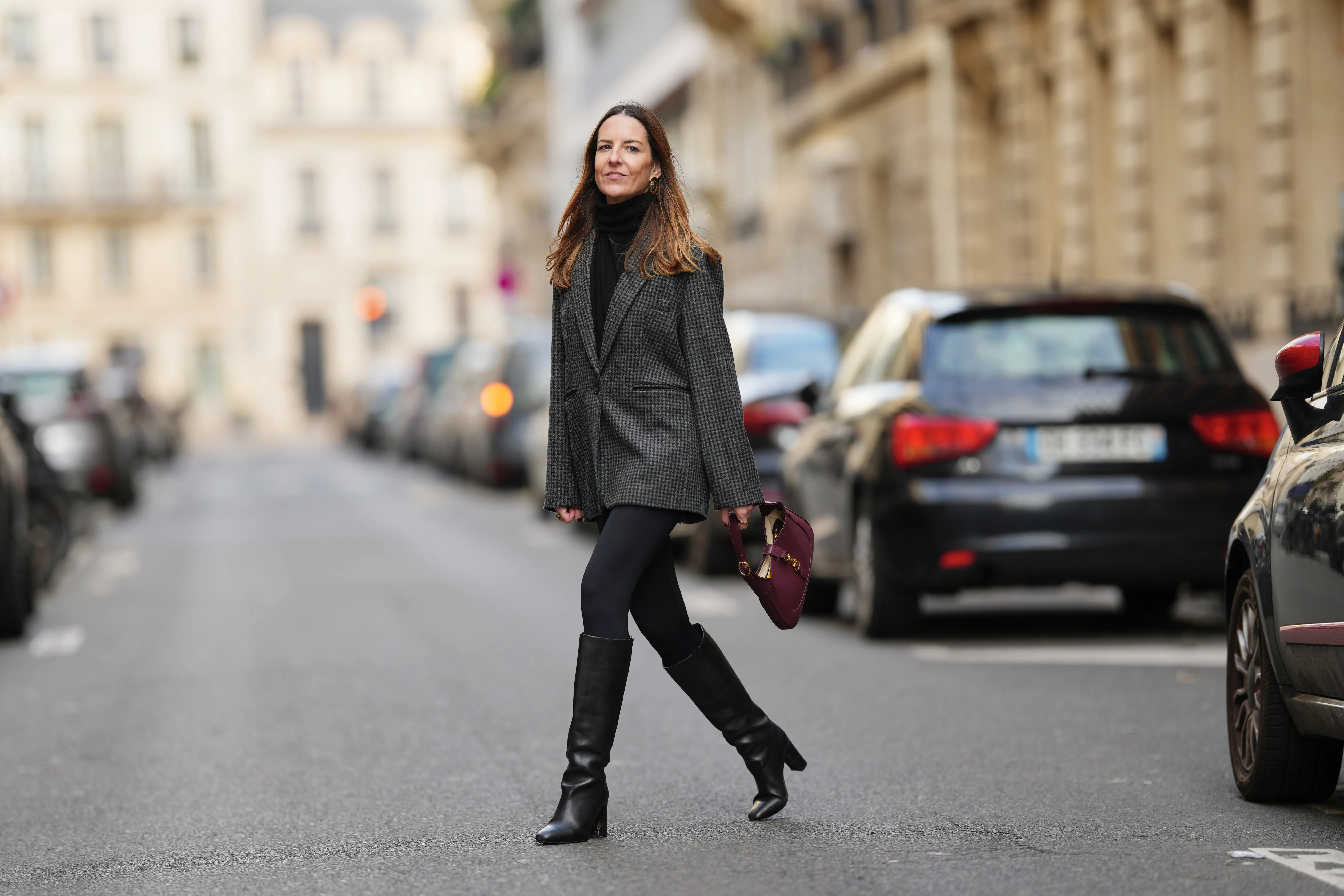 Alba Garavito walks across road wearing gold-tone jewelry by Sempiterno Paris, a black high-neck turtleneck top, a grey houndstooth oversized blazer jacket and black Zara leggings with a fitted silhouette.