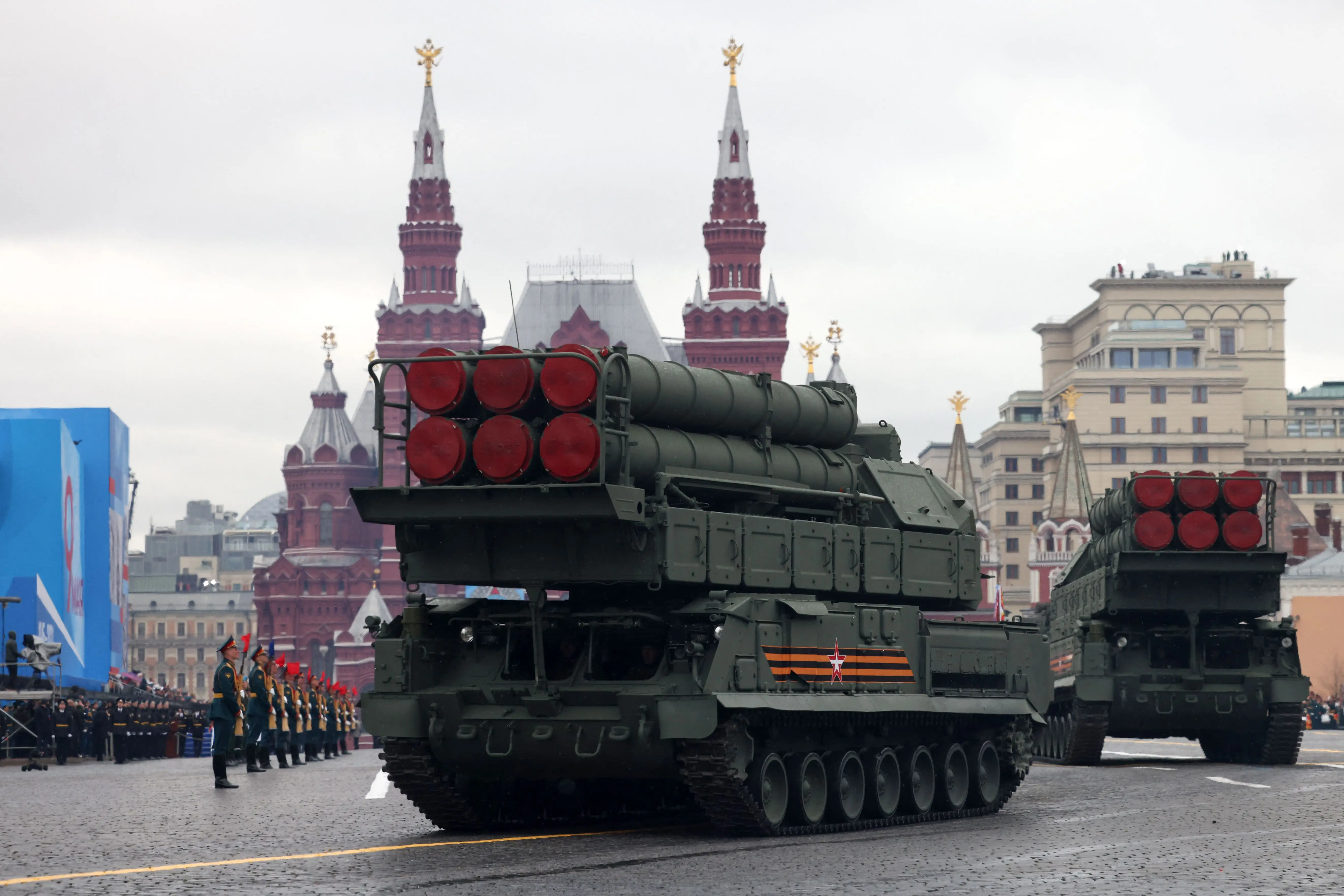 A large green air defense system on wheels with six missiles with red bottoms drives on a street with soldiers beside it under a grey sky and before an elaborate red-brick building