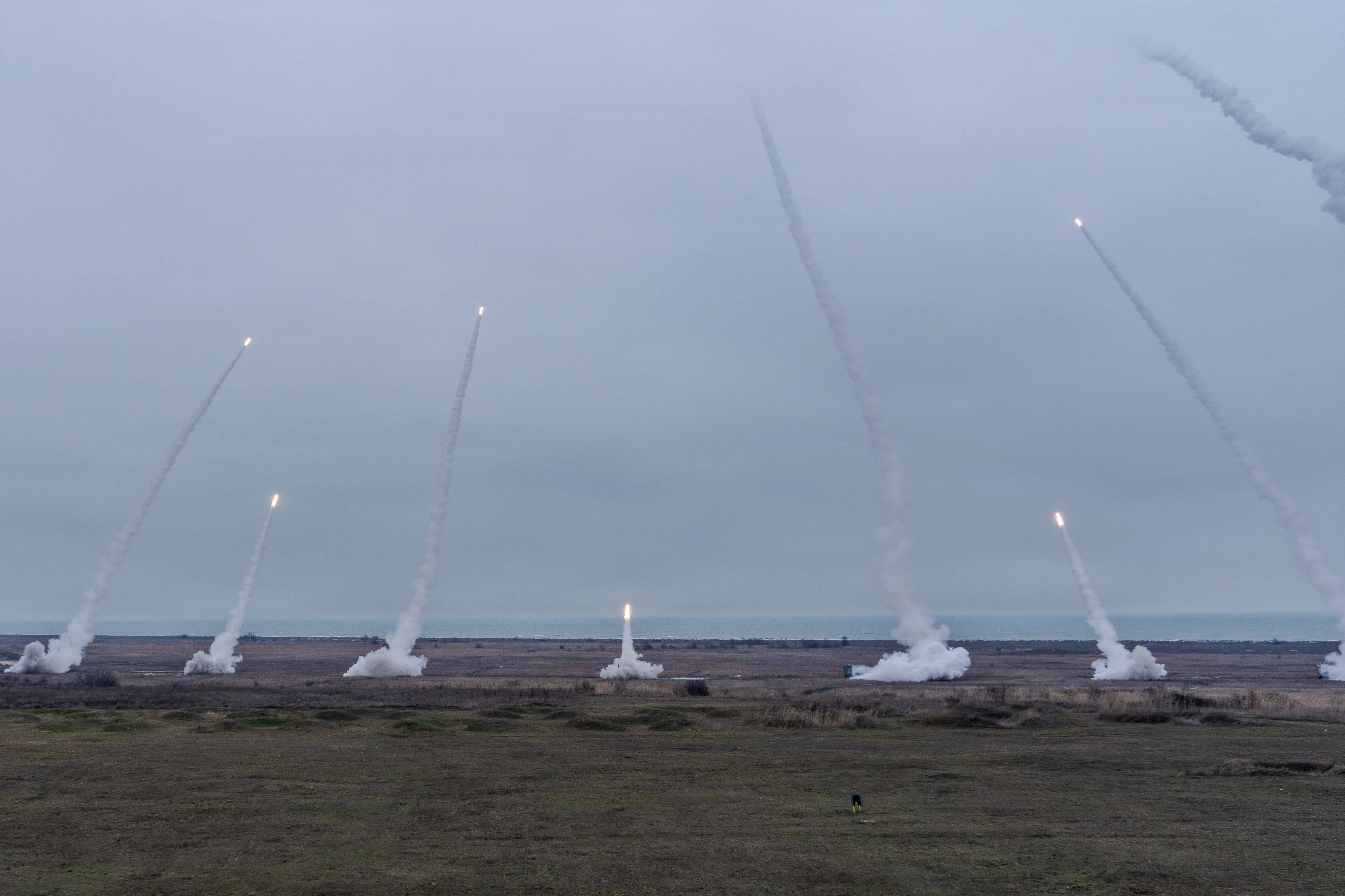 Multiple rocket systems fire from the ground towards an overcast sky.