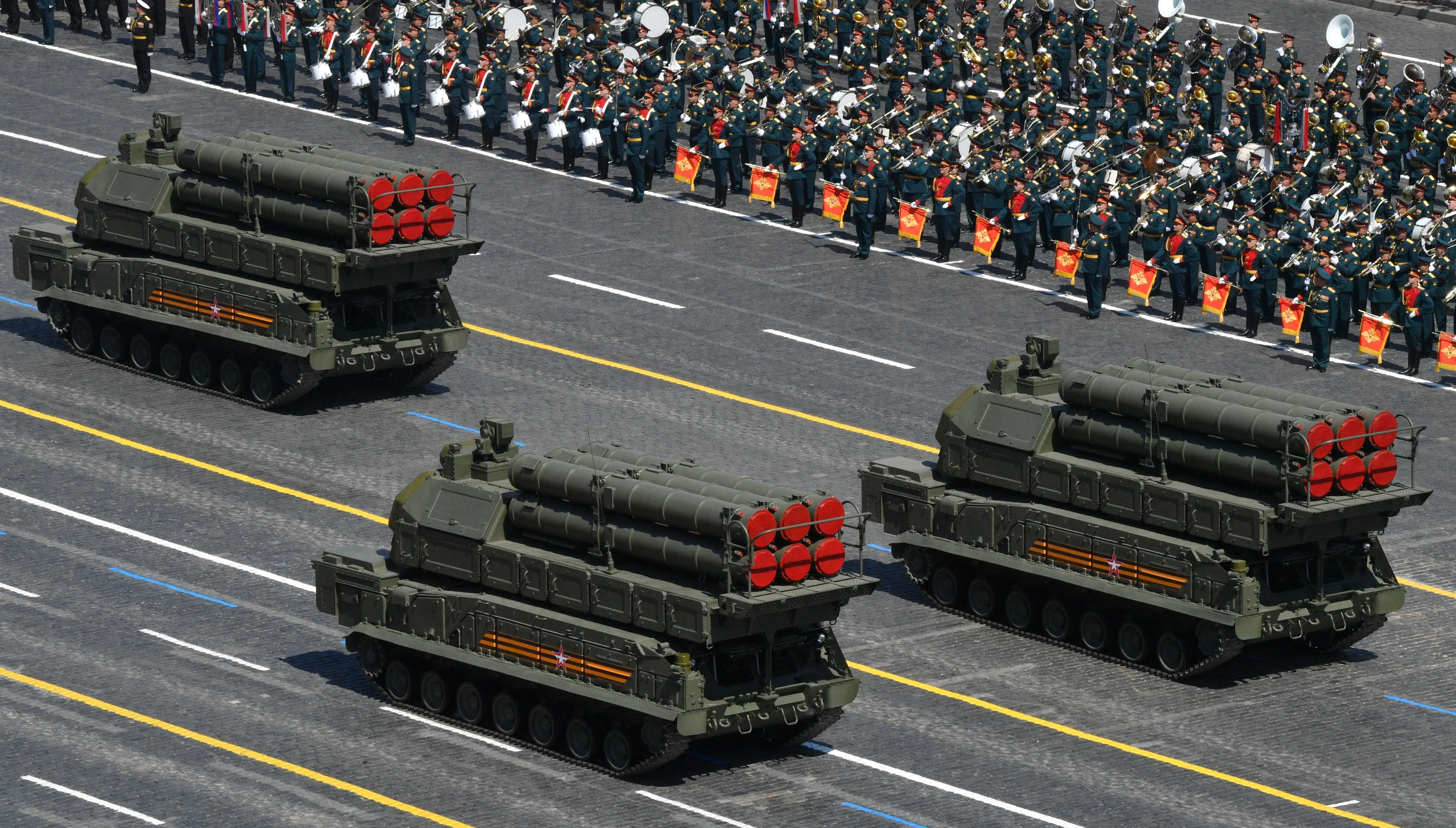 Three large green air defense systems drive on roads with soldiers in green lined up and watching on