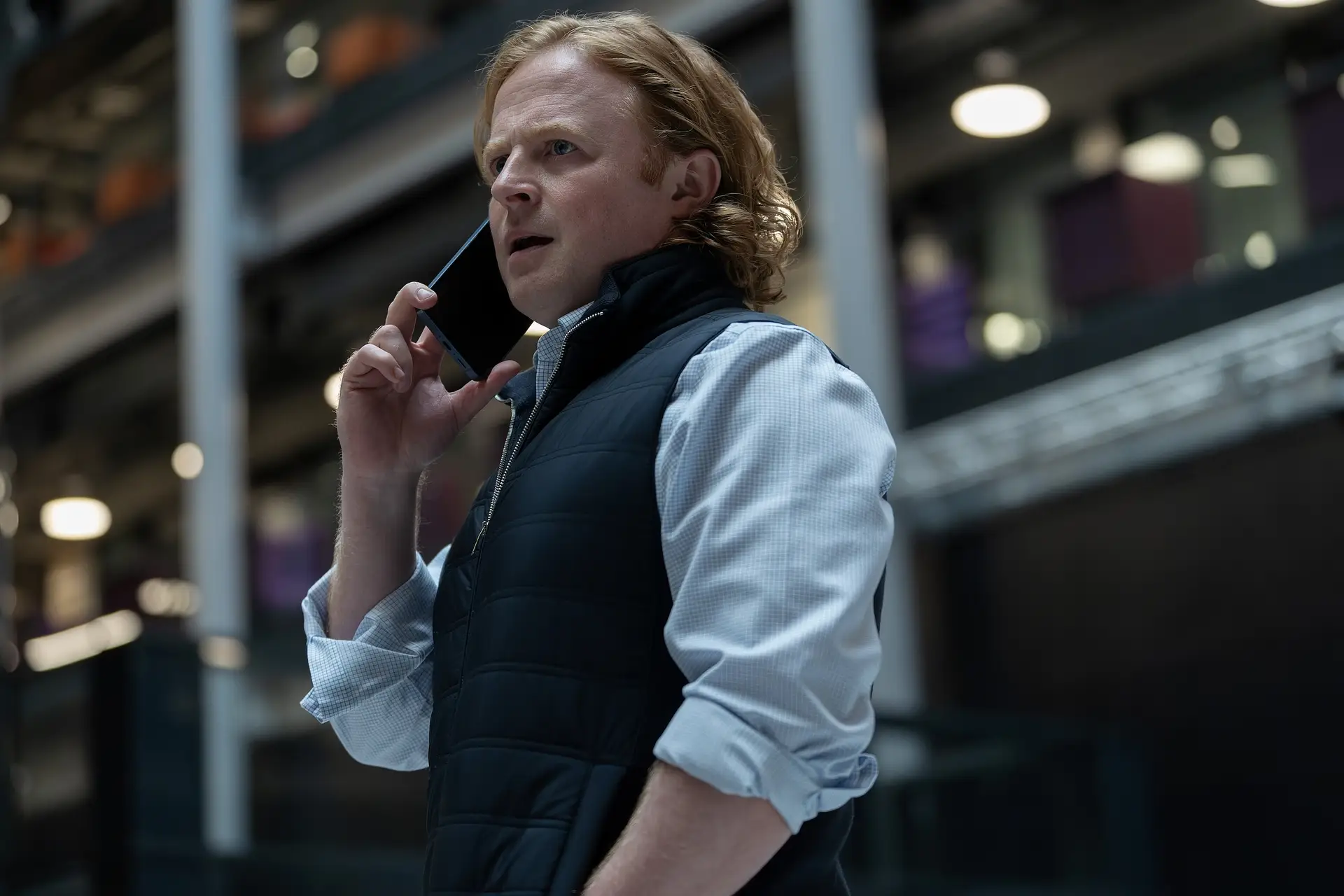 A man wearing a blue checked shirt and navy blue vest takes a phone call.