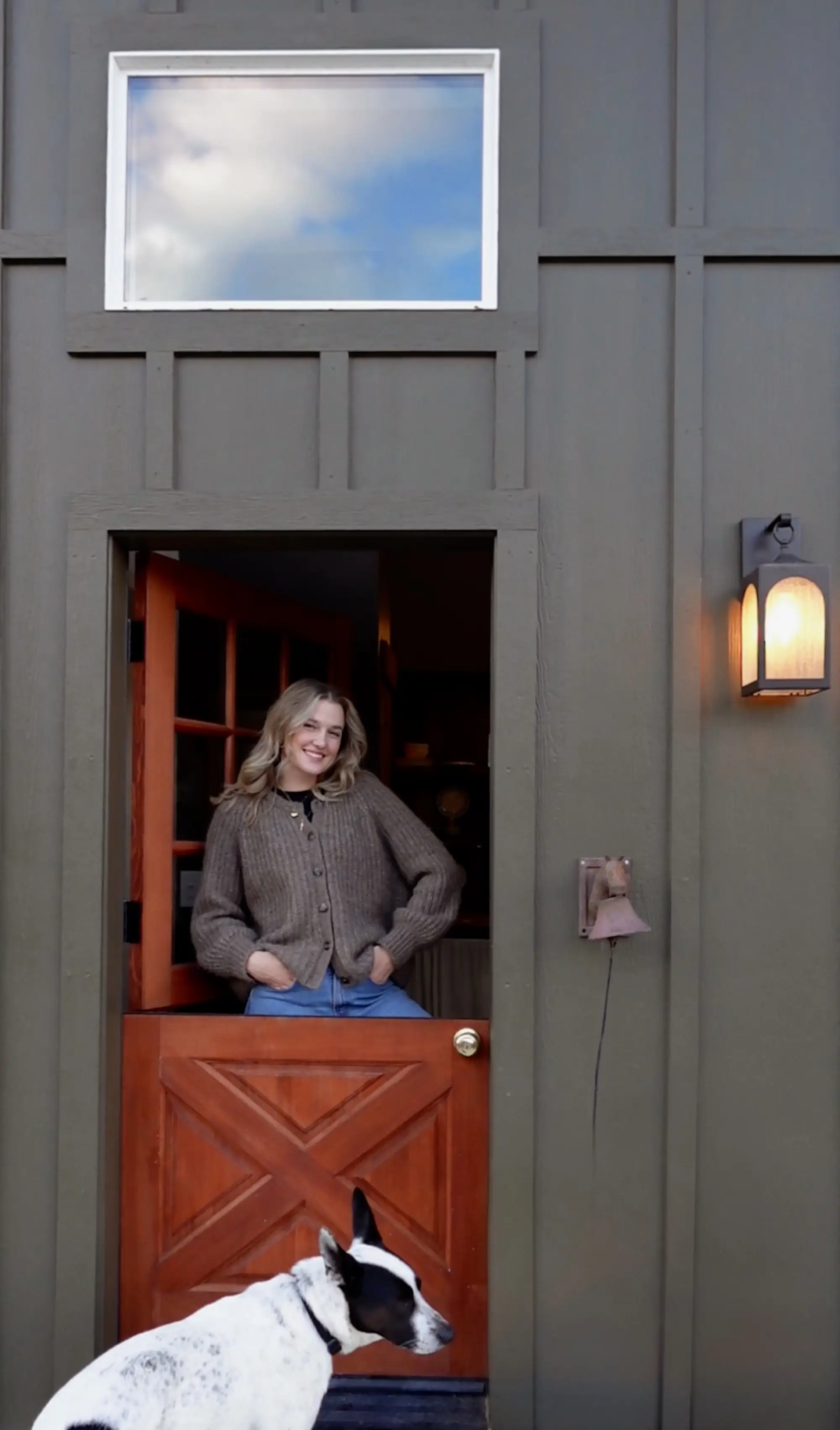 A woman stands in a Dutch door with a dog in front of it.