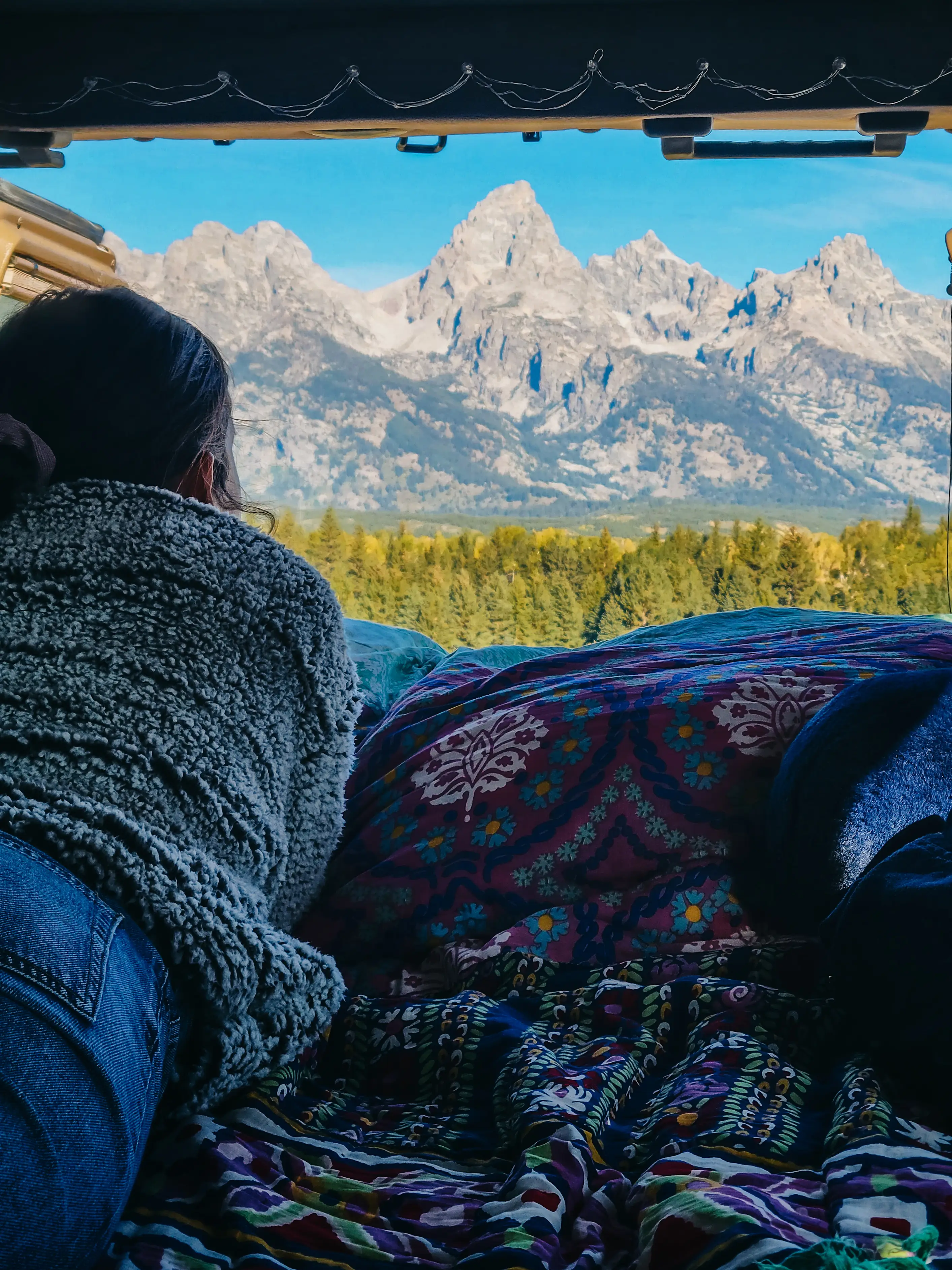 A woman sits in the back of an open van, mountains stand before her.