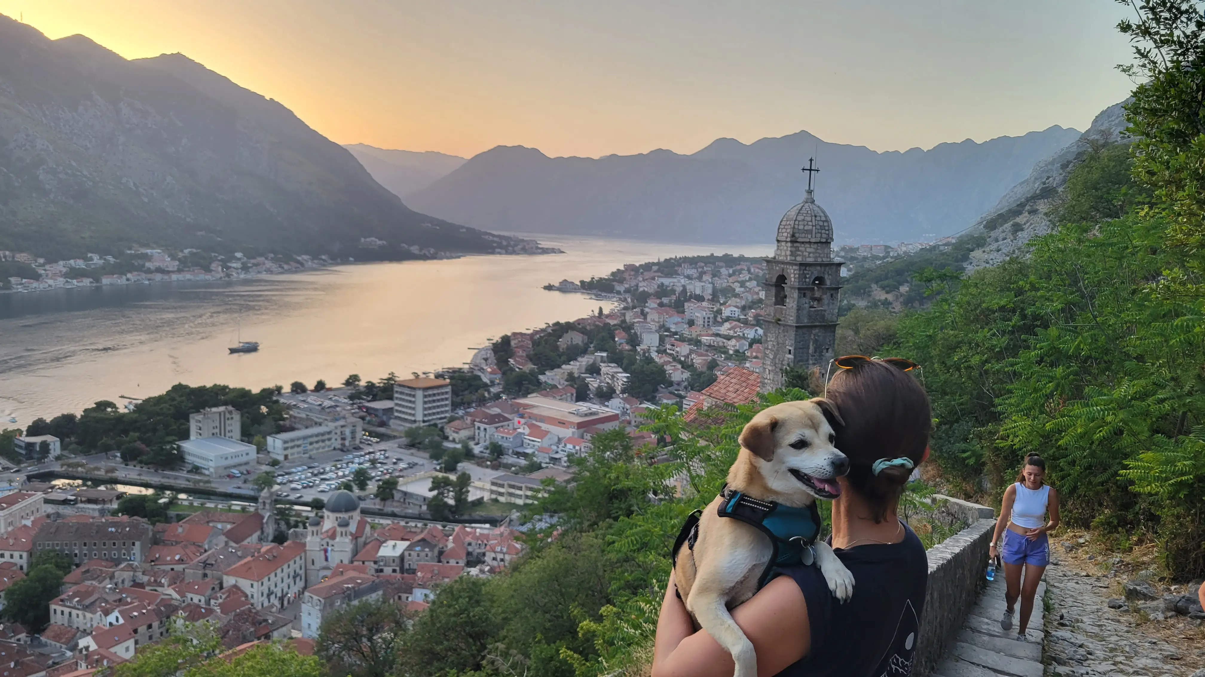 A woman and her dog stand on a walking trail, sitting high above a city in Montenegro.