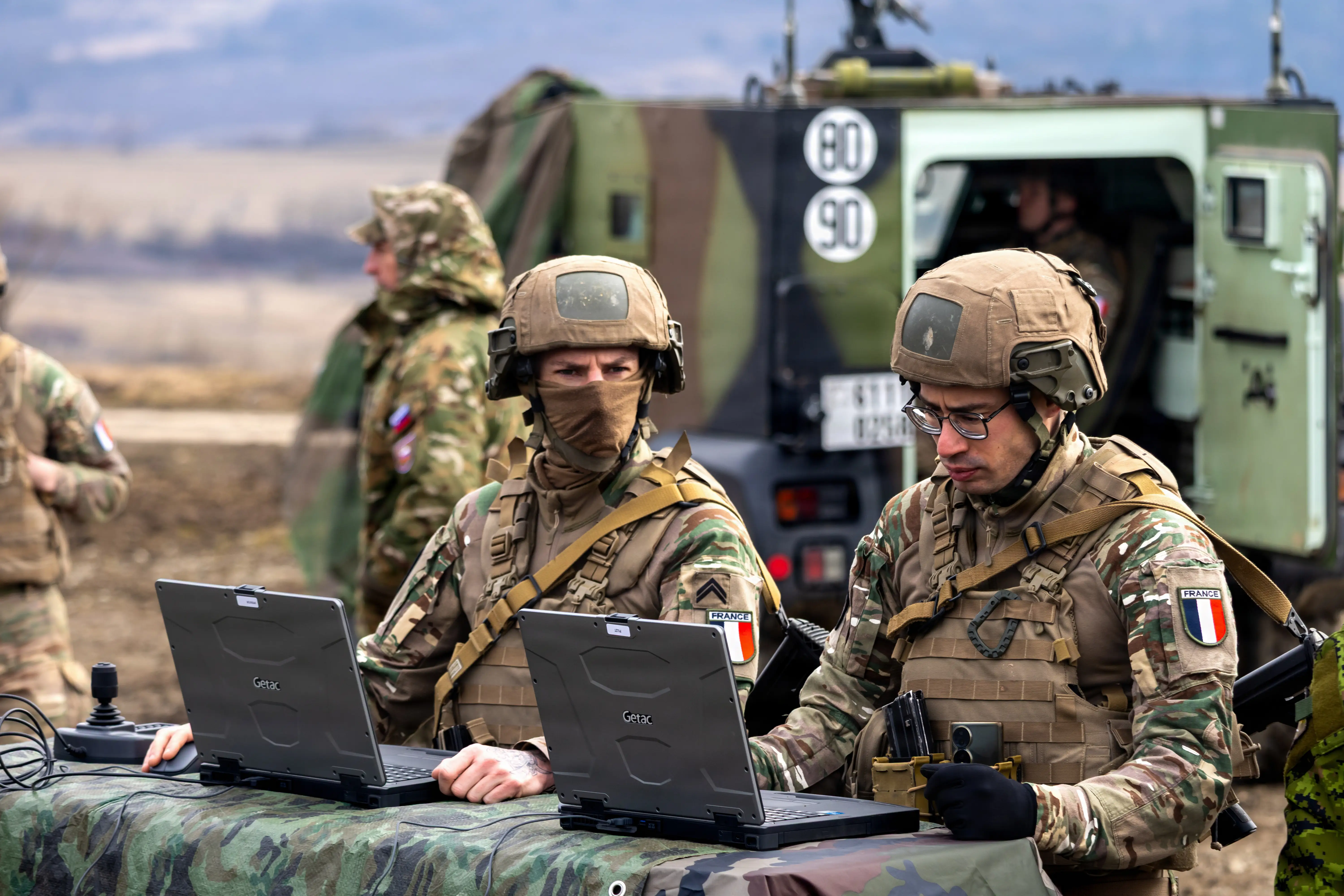 Two soldiers sit at a table working on laptops.