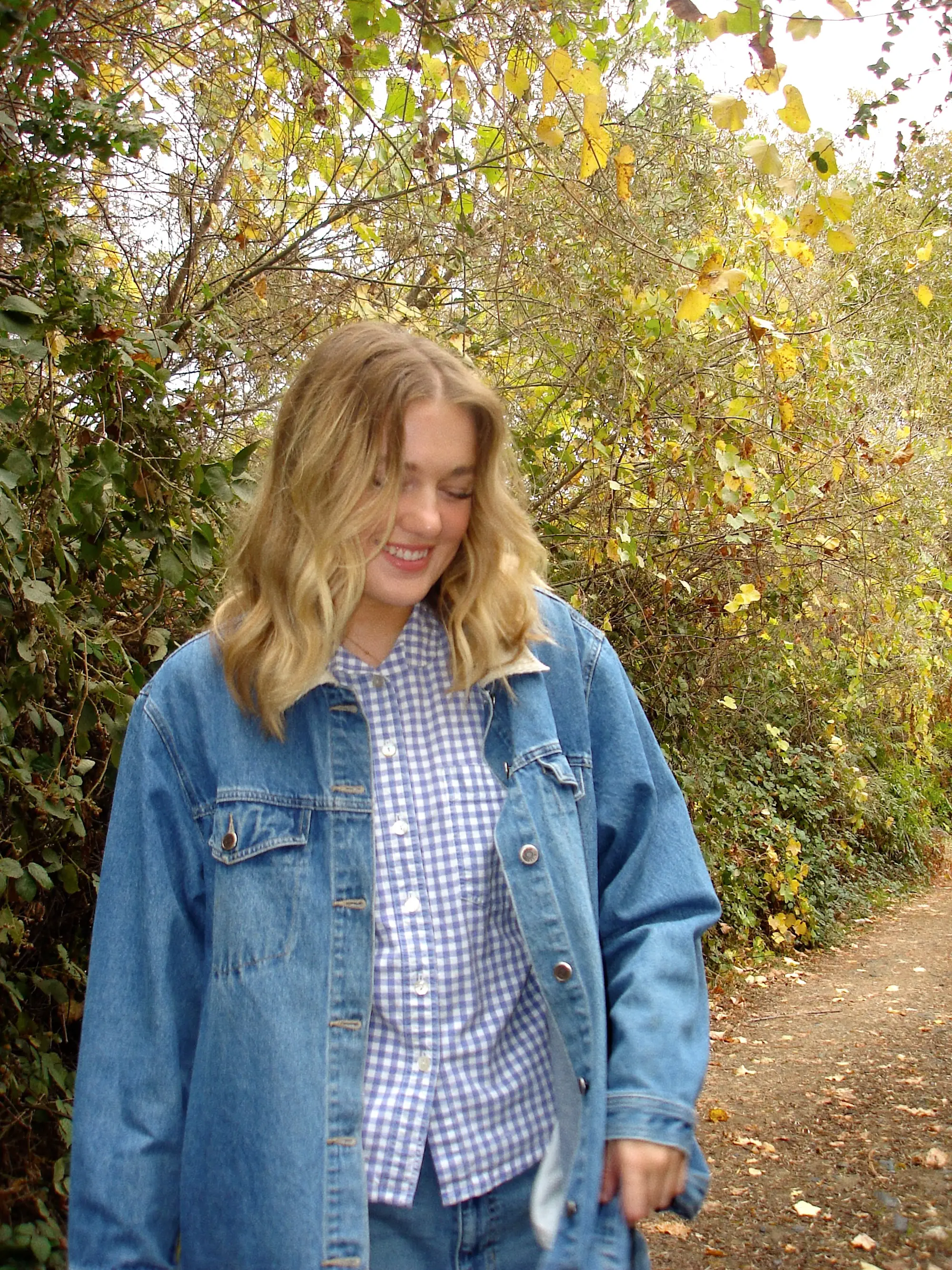 A blond woman smiles and looks down with trees behind her.