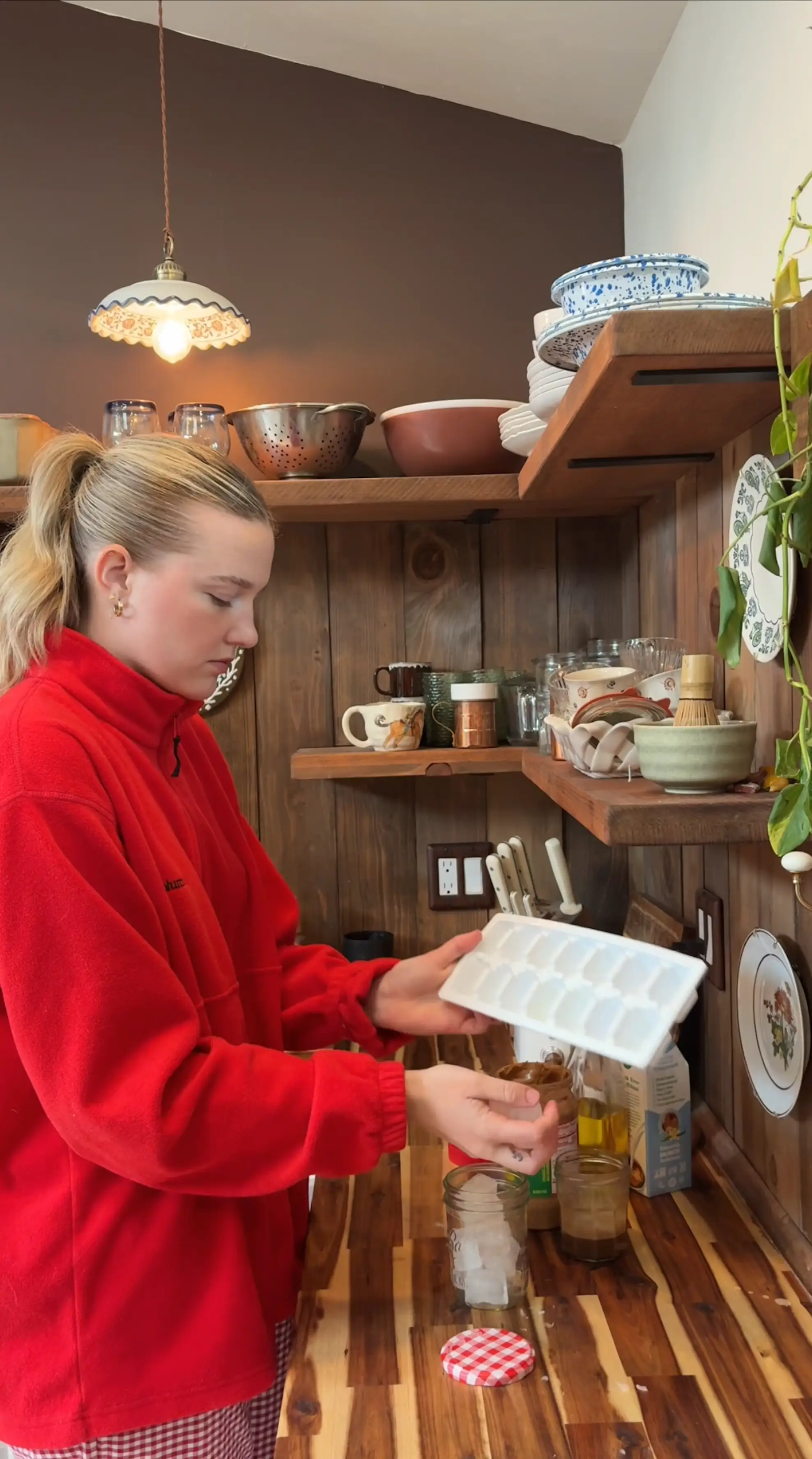 A woman makes a latte in her kitchen that has wooden backsplash and counters, as well as open shelving.