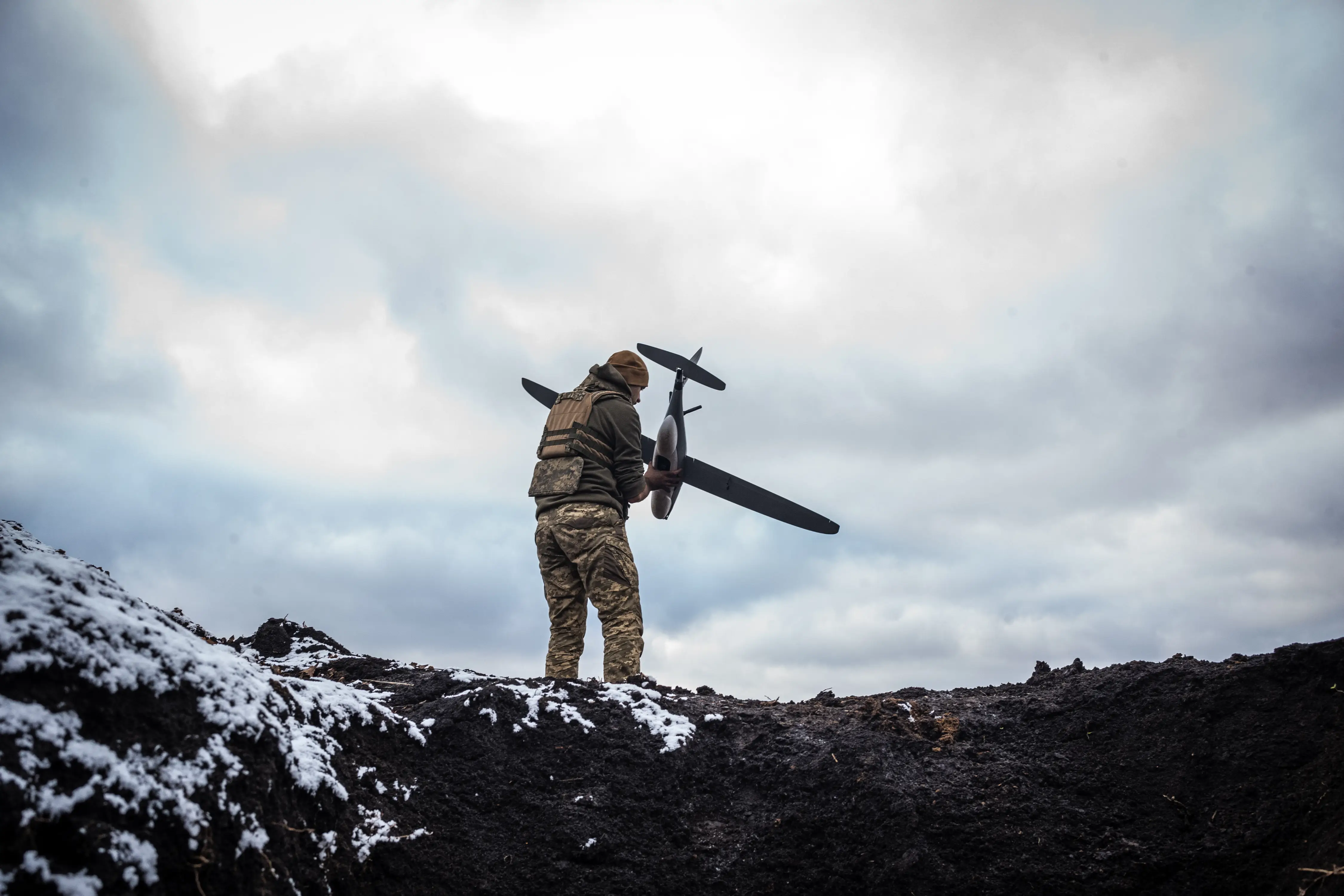 A man in camouflage gear stands with his back turned and holding a small drone while standing on snowy and muddy ground under a cloudy sky