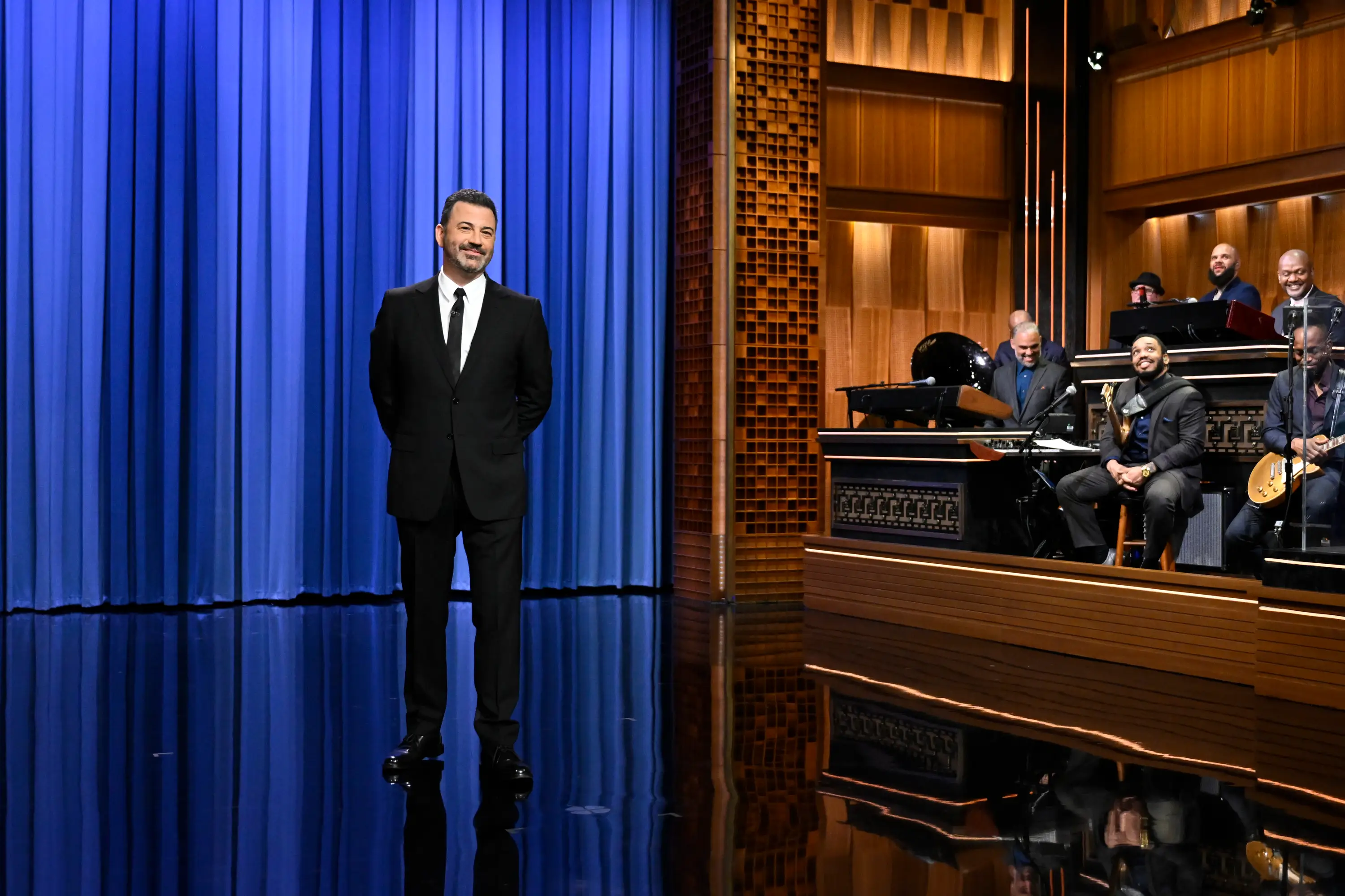 Jimmy Kimmel is standing on stage in a black suit with a black tie. He is in front of a navy blue drape.