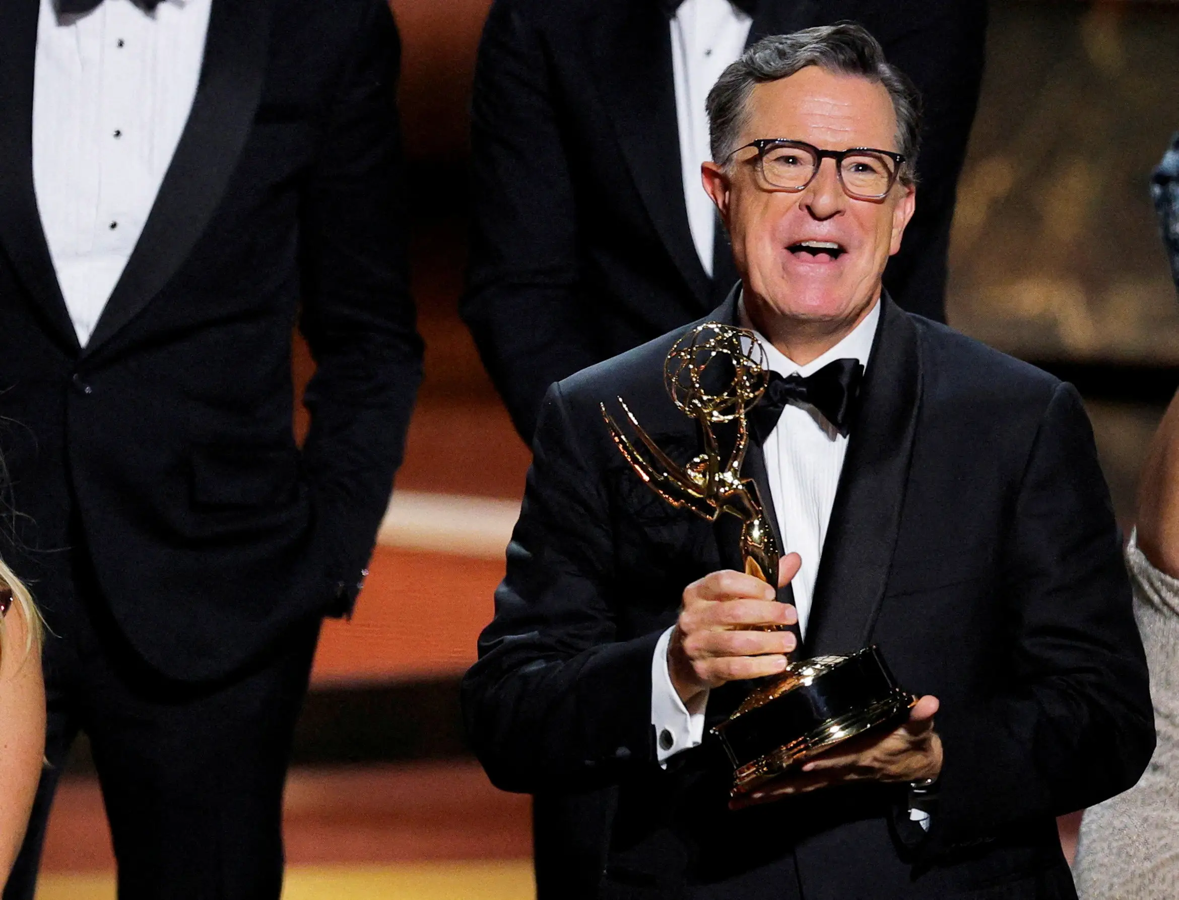 Stephen Colbert is holding an Emmy Award trophy. He is wearing a black suit with a black bowtie.
