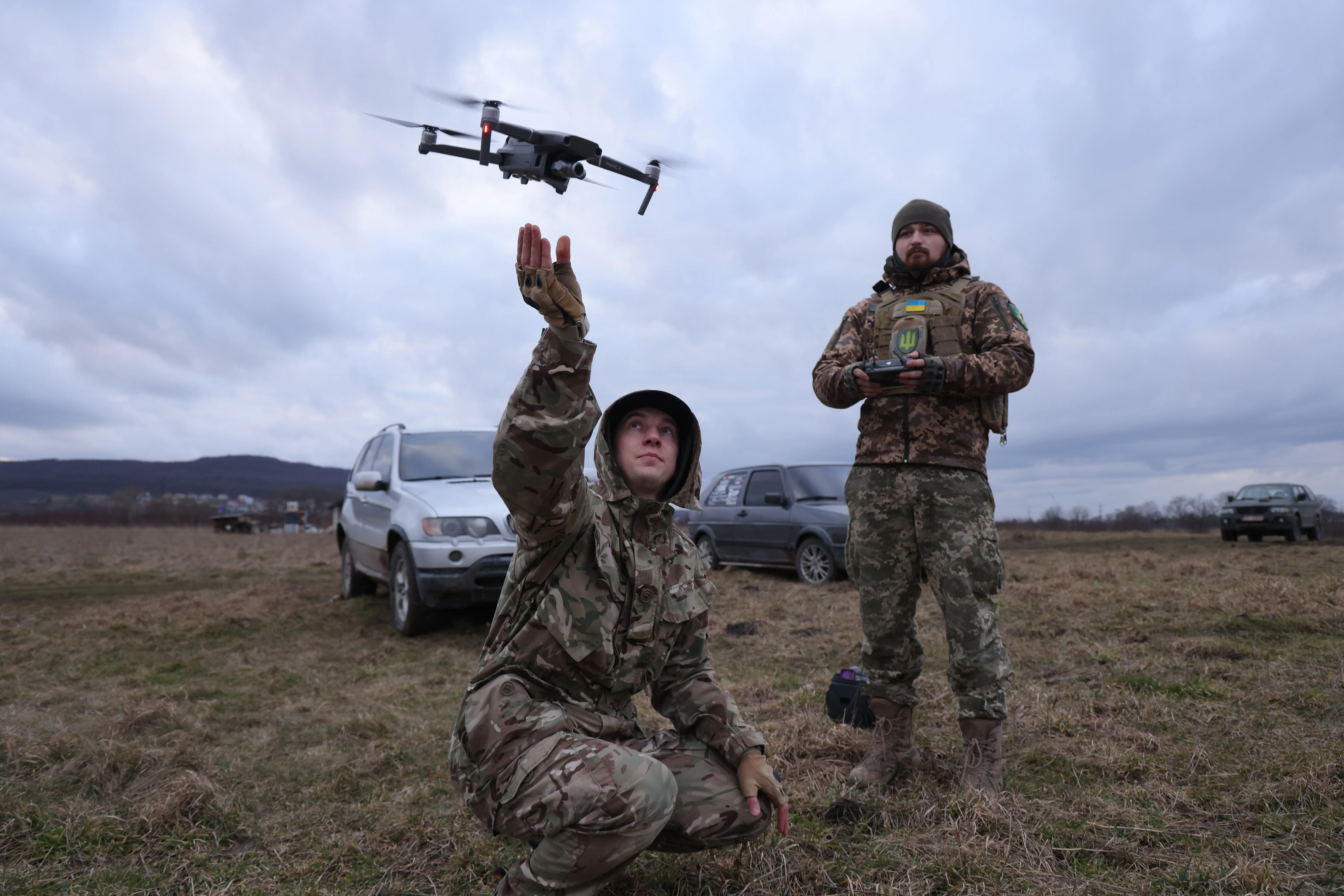 A figure in camouflage gear squats with their arm up and a small drone hovering above him, with another figure in camouflage standing behind and holding a controller, under a grey sky and on grass and with two cars, light and dark grey, behind them