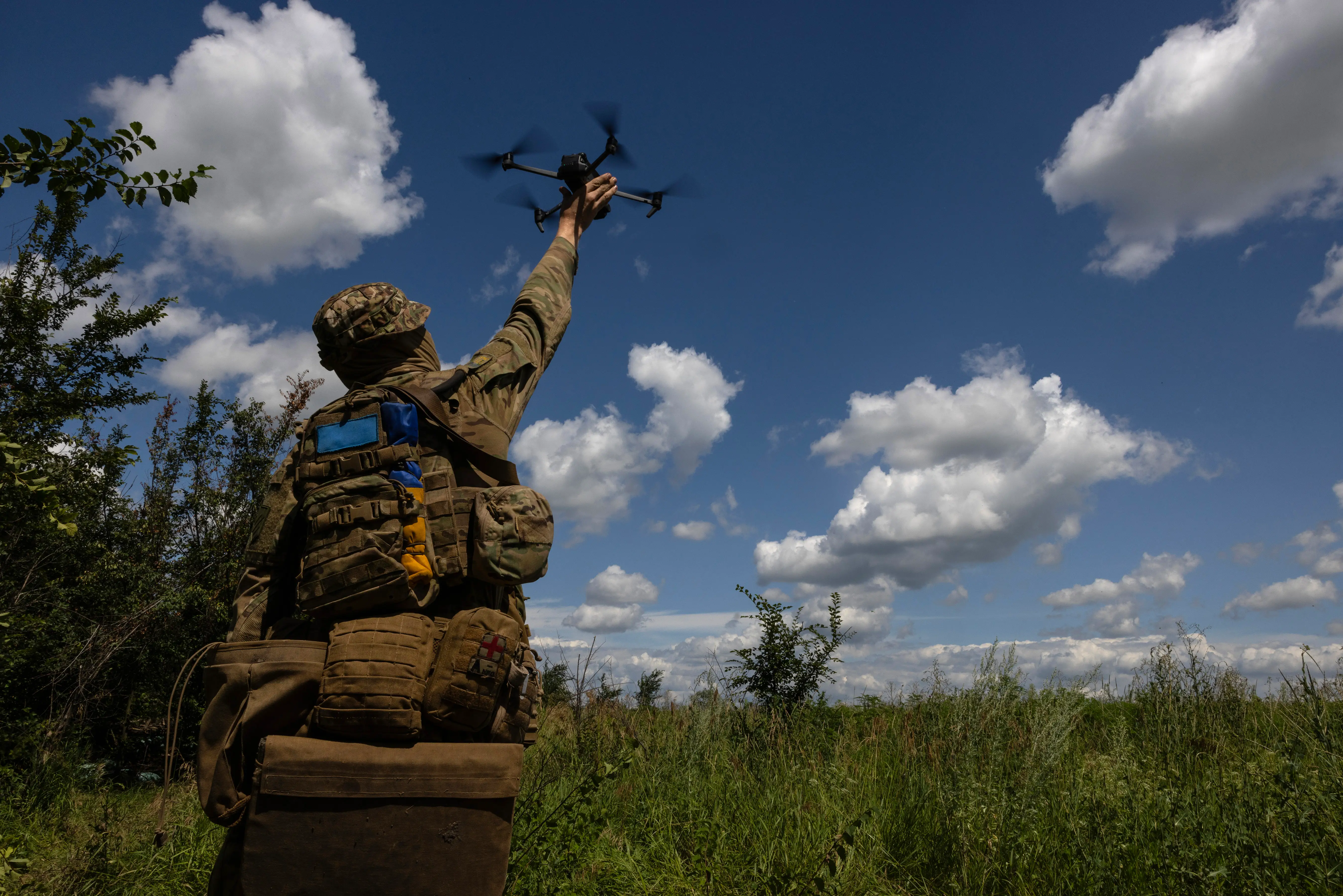 A figure in camouflage gear and with their back turned holds an arm up holding a small black drone under a blue cloudy sk,y and on shubbery