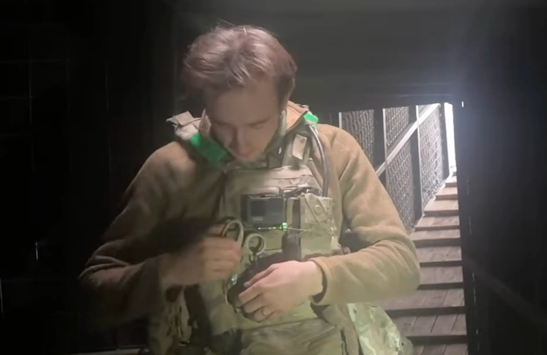 A man in khaki gear stands in a dark space with stairs leading up to light behind him, pulling out scissors from a pouch at his chest