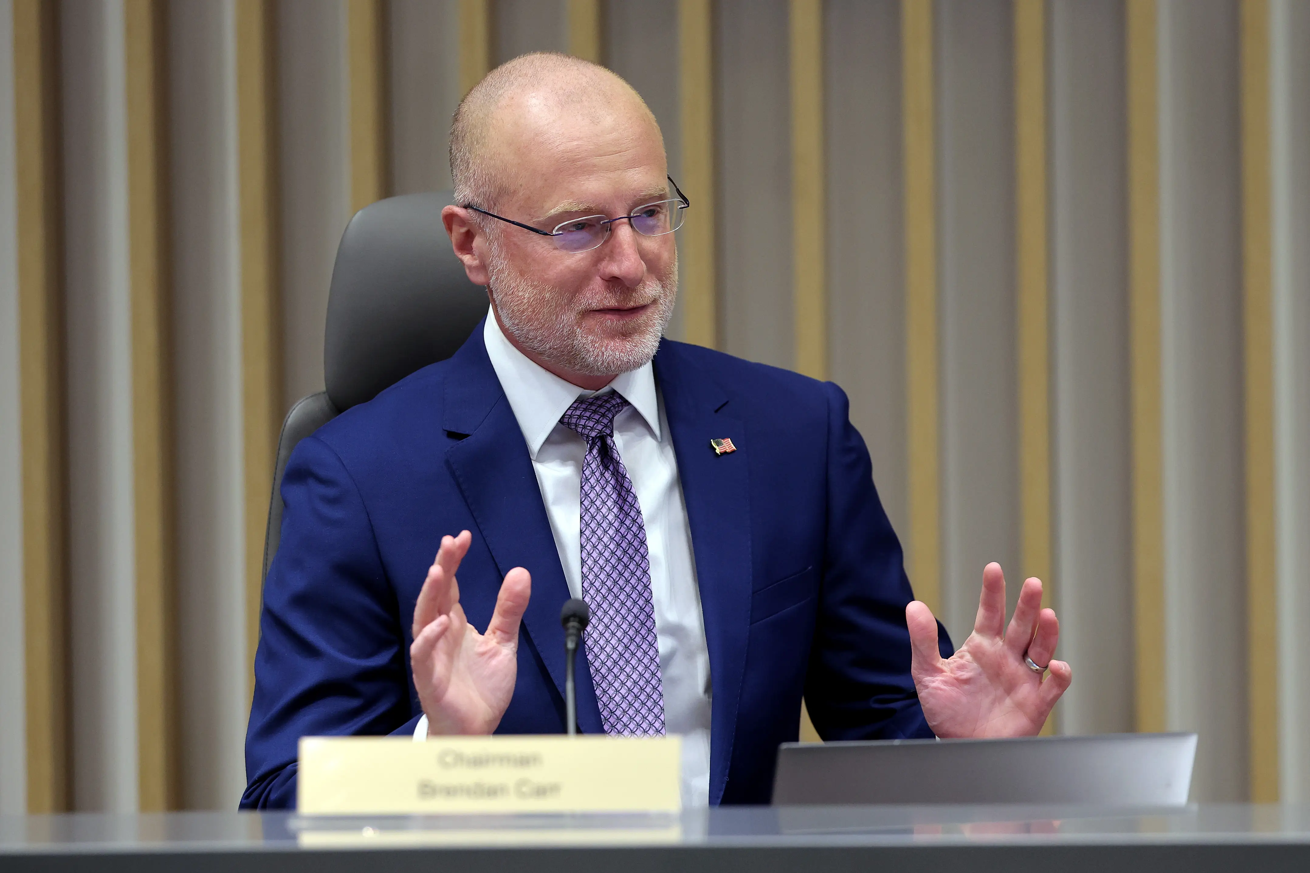 FCC Chair Brendan Carr sits behind a desk with his hands up.