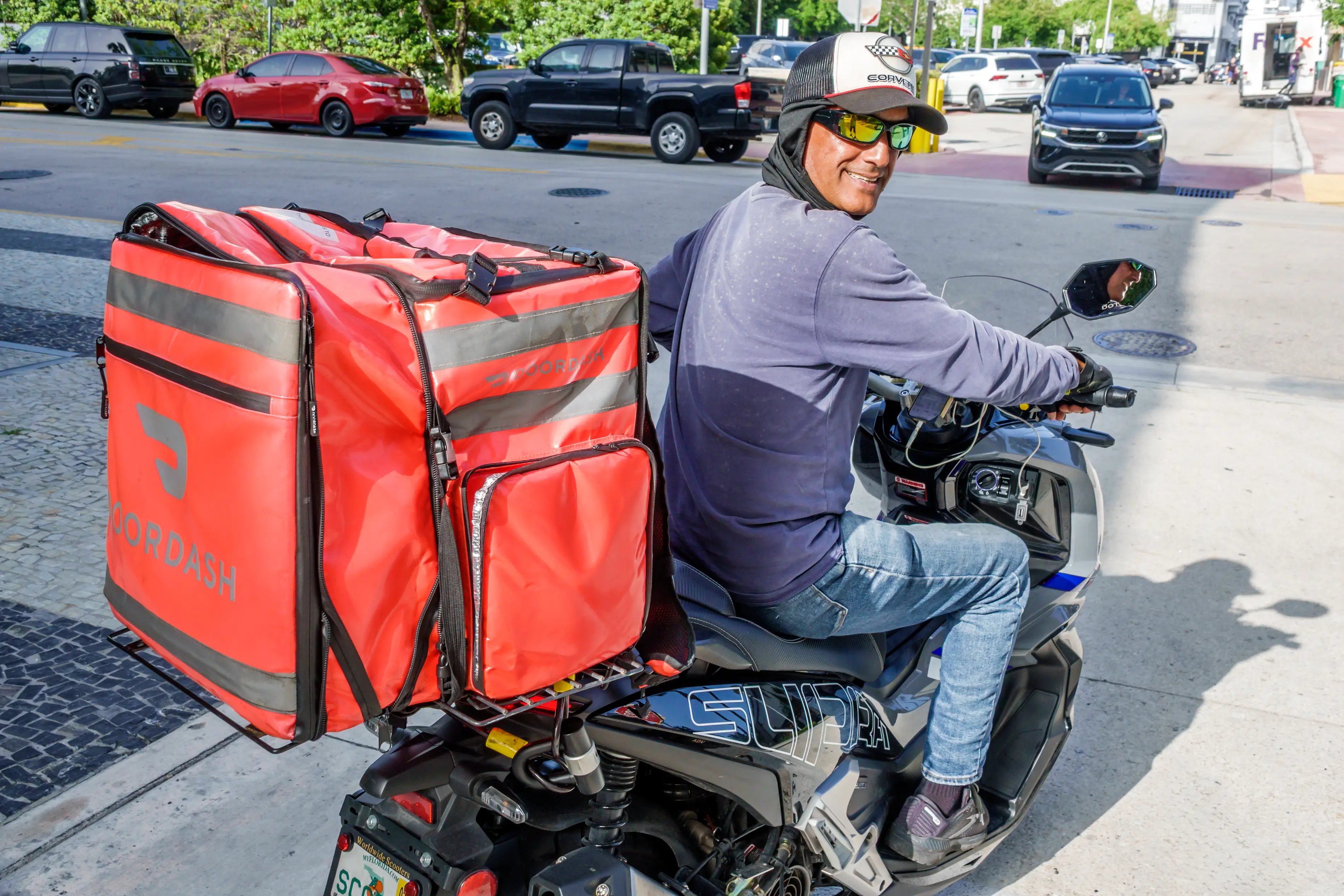 A DoorDash delivery worker drives a motorcycle with a red padded bag mounted on the back as he looks back at the photographer and smiles while wearing sunglasses.