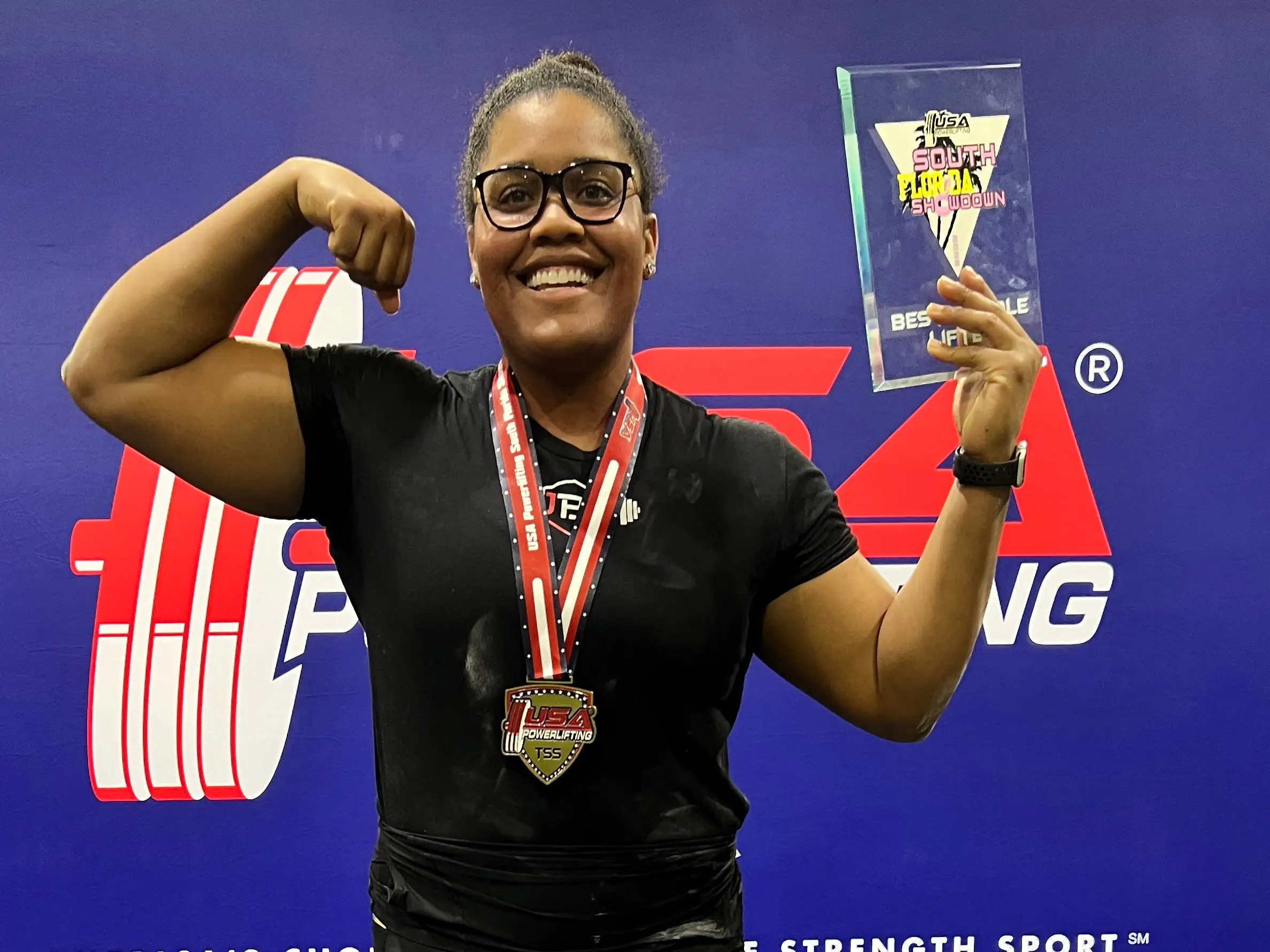 a woman standing on a podium with a medal and award during a powerlifting meet.