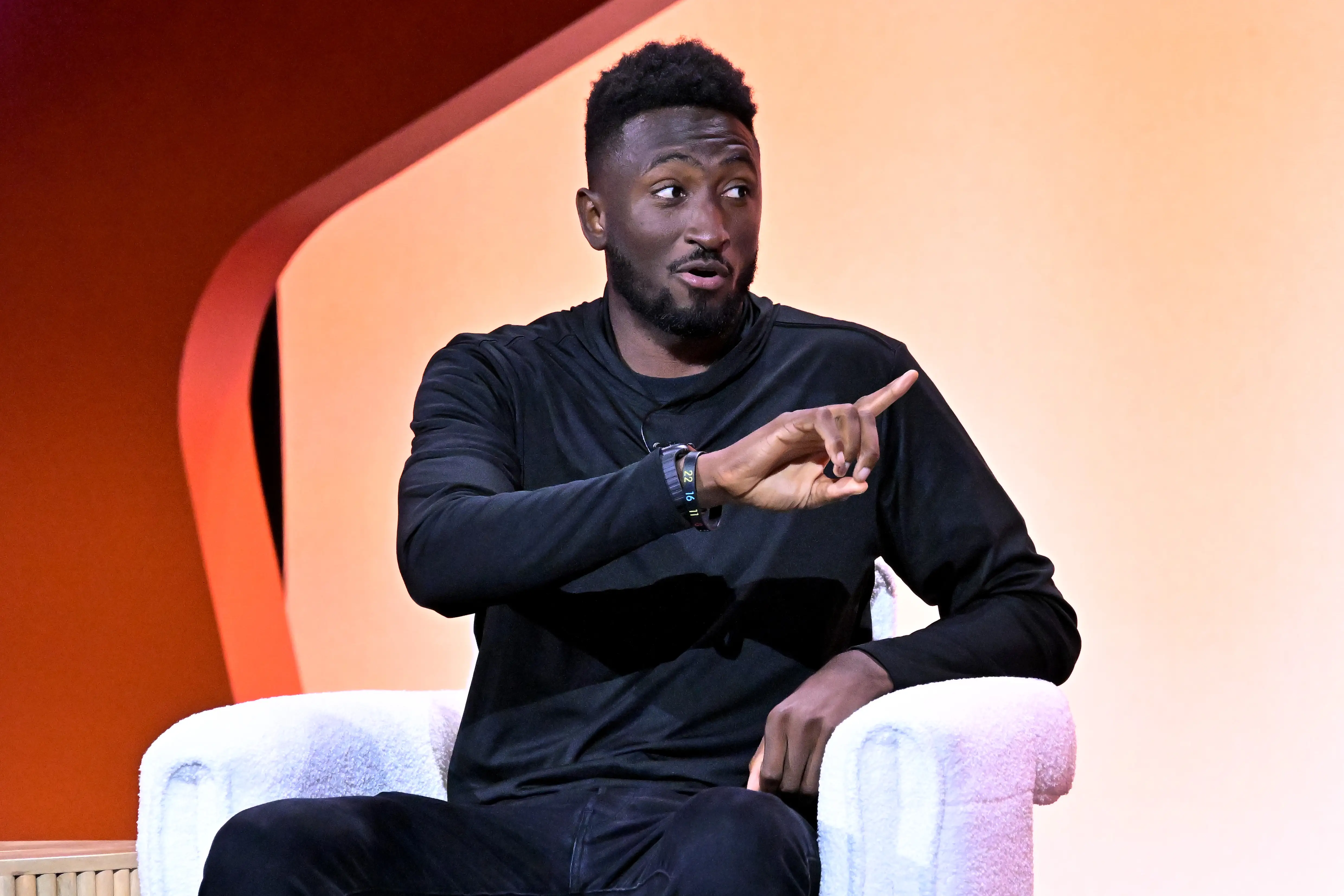 Marques Brownlee, dressed in a long-sleeve black shirt and black pants, sits on a white chair. He is pointing to his left.