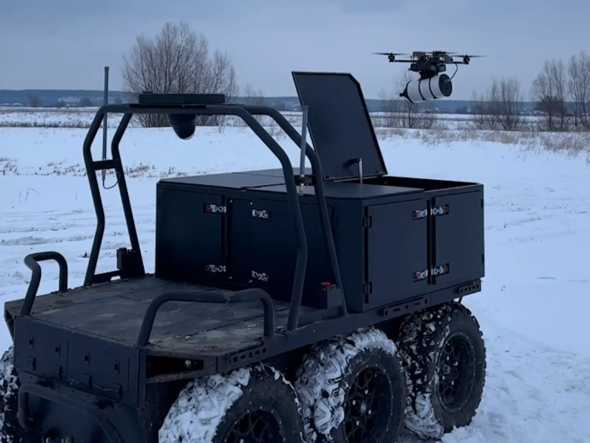 A black ground robot on six wheels on snow with a drone flying above it under a grey sky
