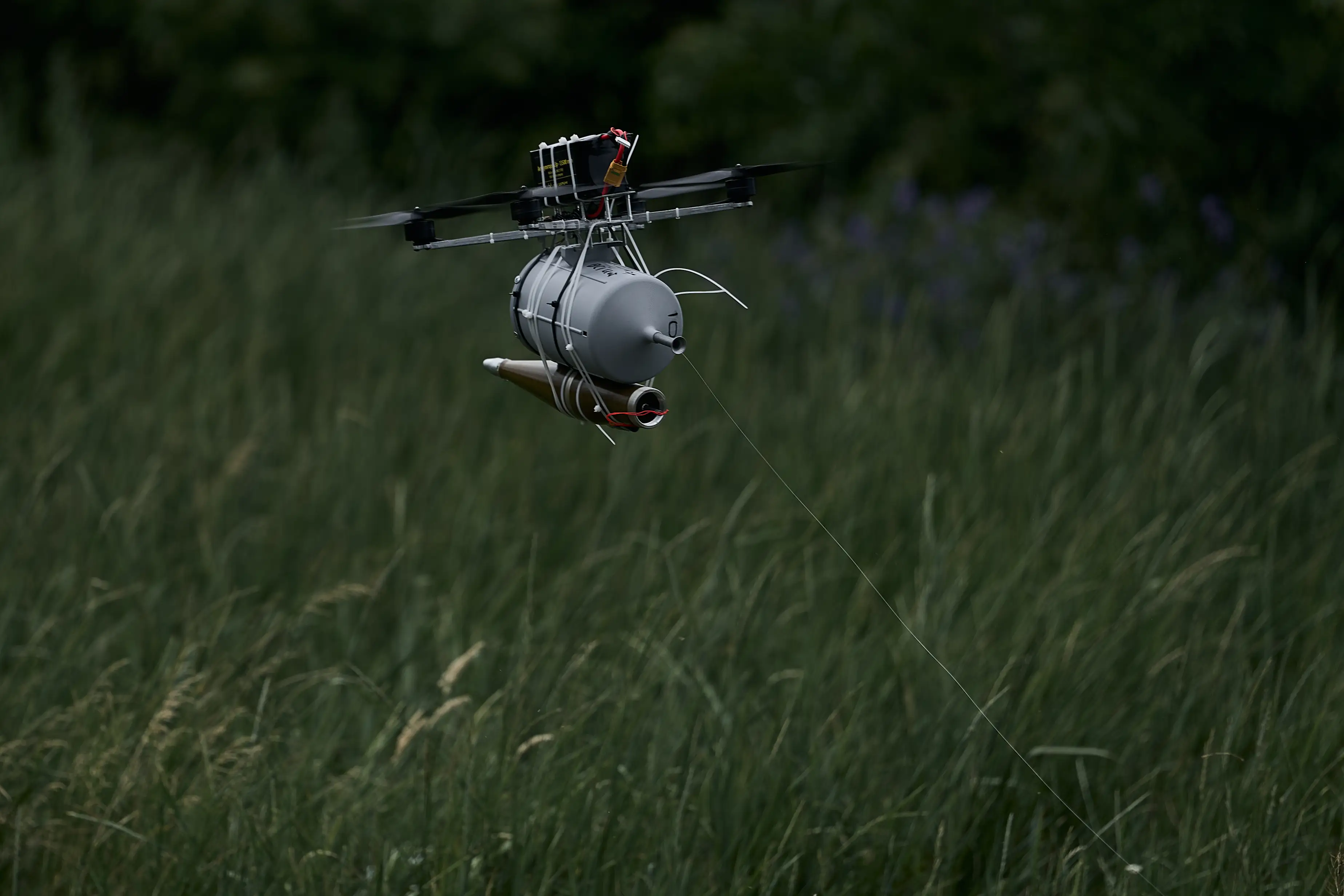 a small black drone above long grass with a grey cylinder underneath it and a long thin cable coming out of it