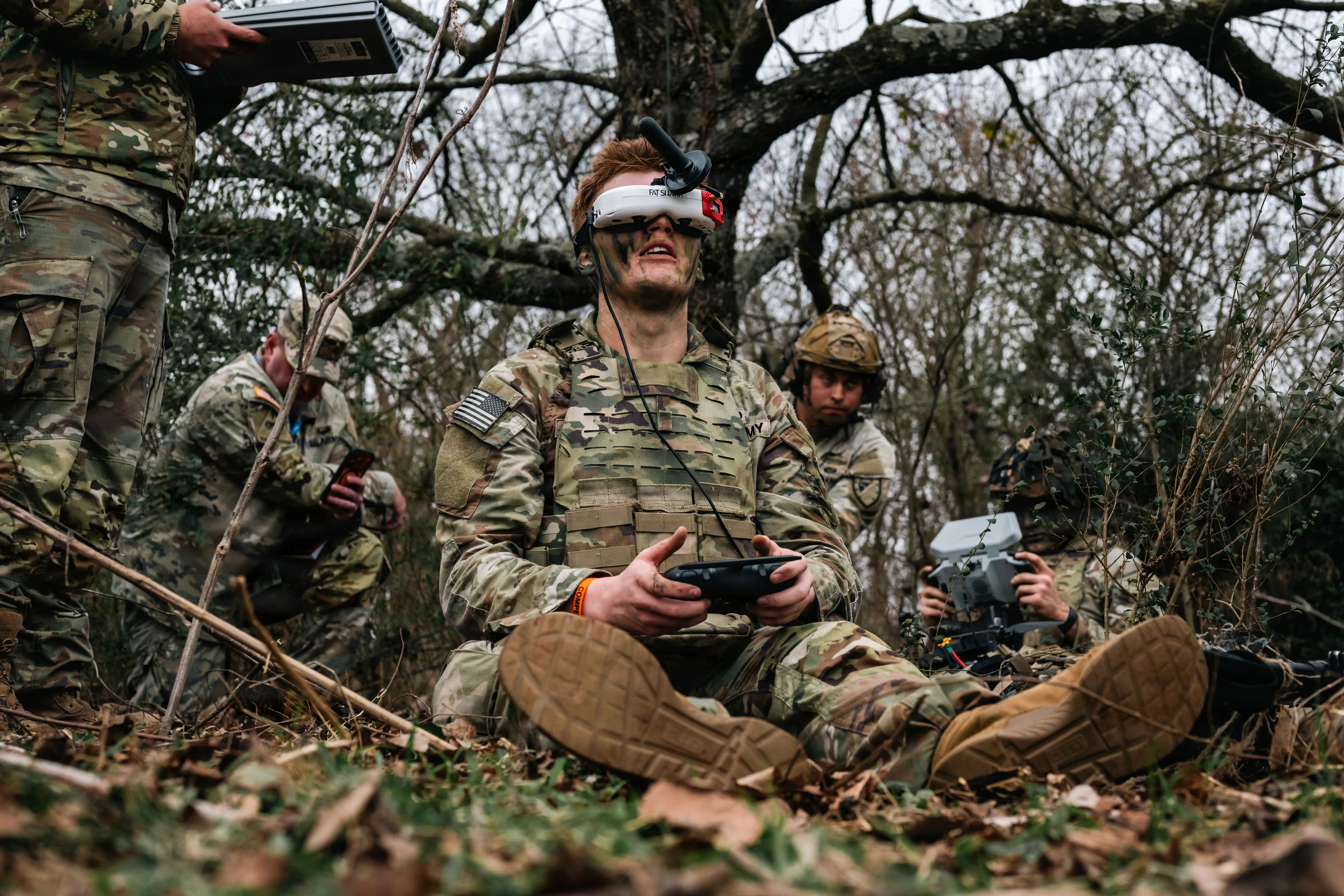 A soldier sits on the ground wearing a headset and holding a controller. Behind him, various soldiers crouch, sit, and stand. There is a barren tree line in the background.