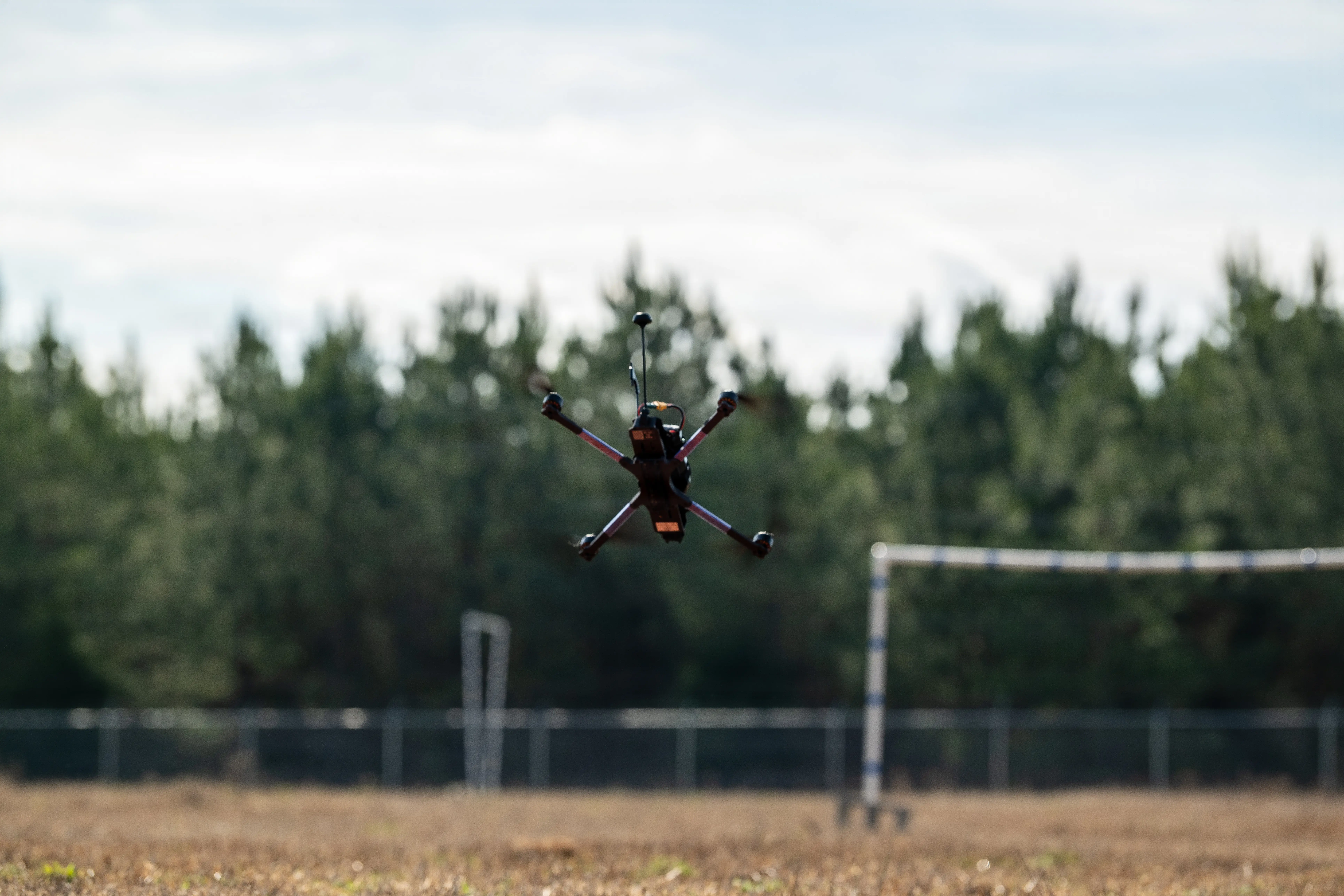 A quadcopter drone flies on a field with trees in the background.