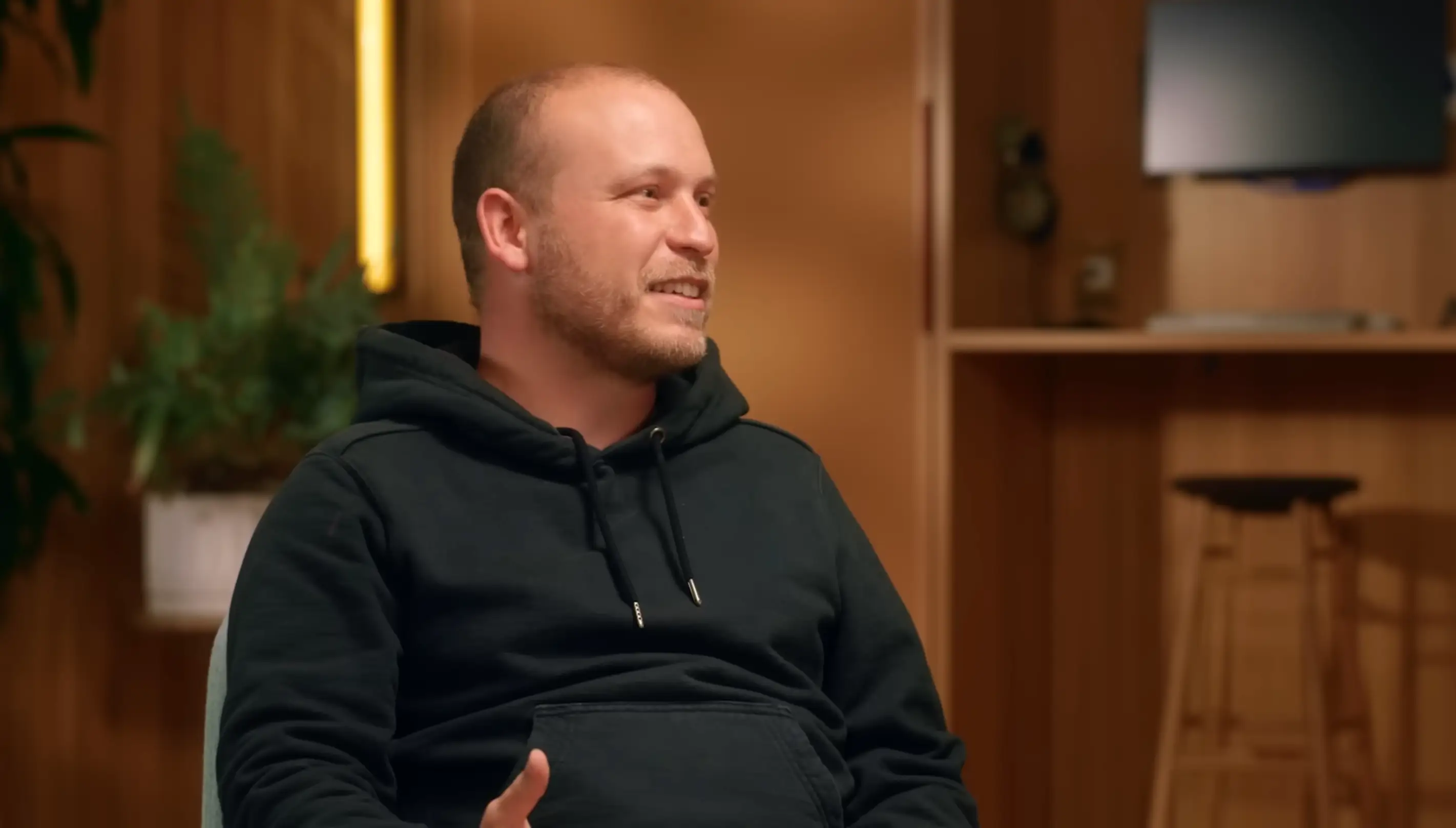 Boris Cherny, a Claude executive, sits in a studio during an interview for Anthropic. He is wearing a black hoodie. The room is filled with light-colored wood wall panels and a few plants.