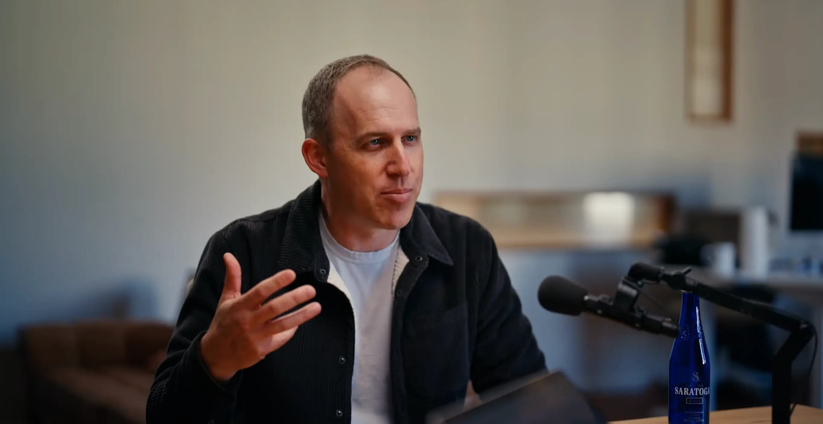 Bret Taylor, OpenAI's board chair, is sitting in front of a podcast mic. He is wearing a black button-down collar shirt with a white undershirt.