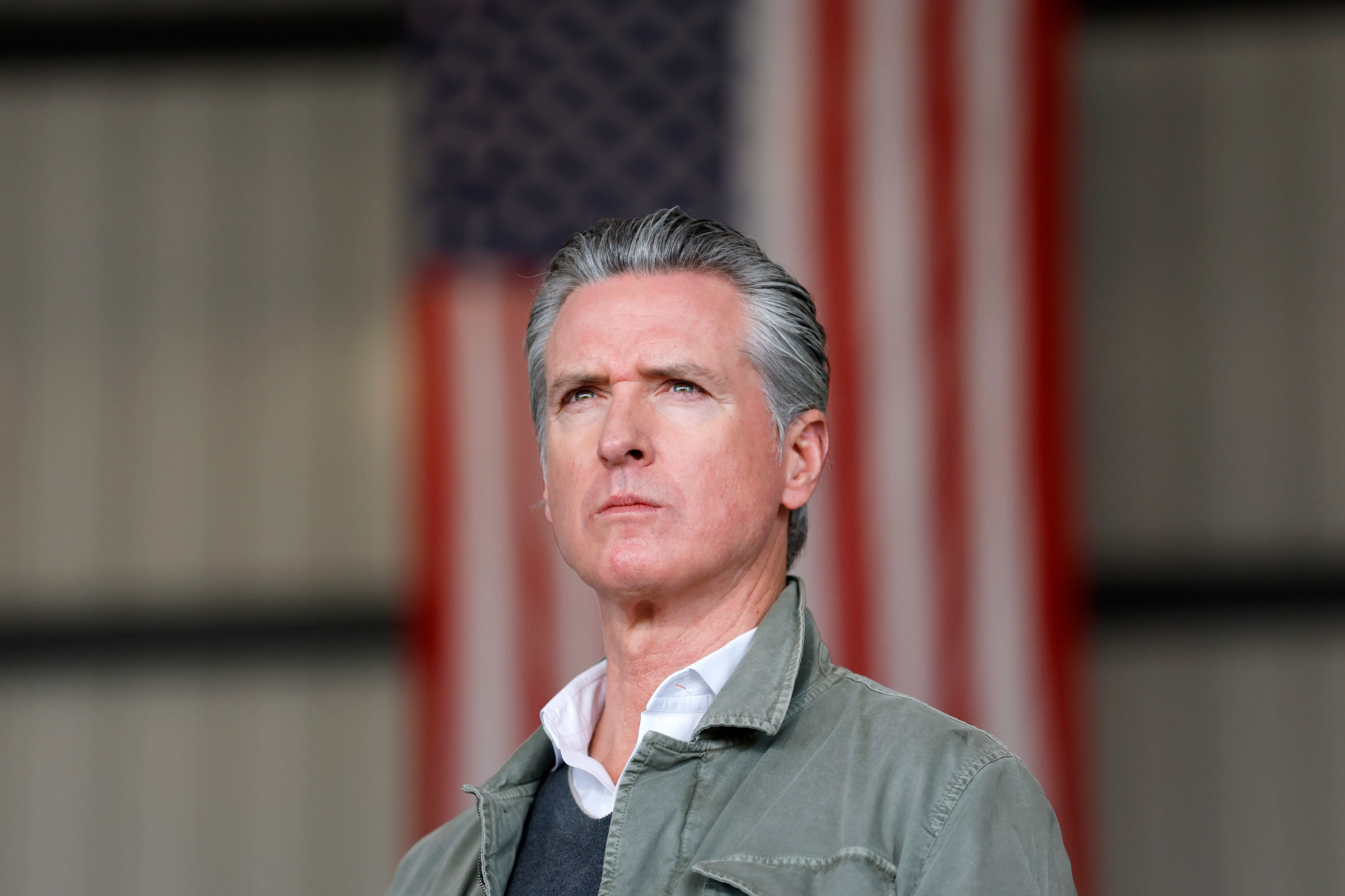 Gavin Newsom in a green jacket and an American flag in the background.