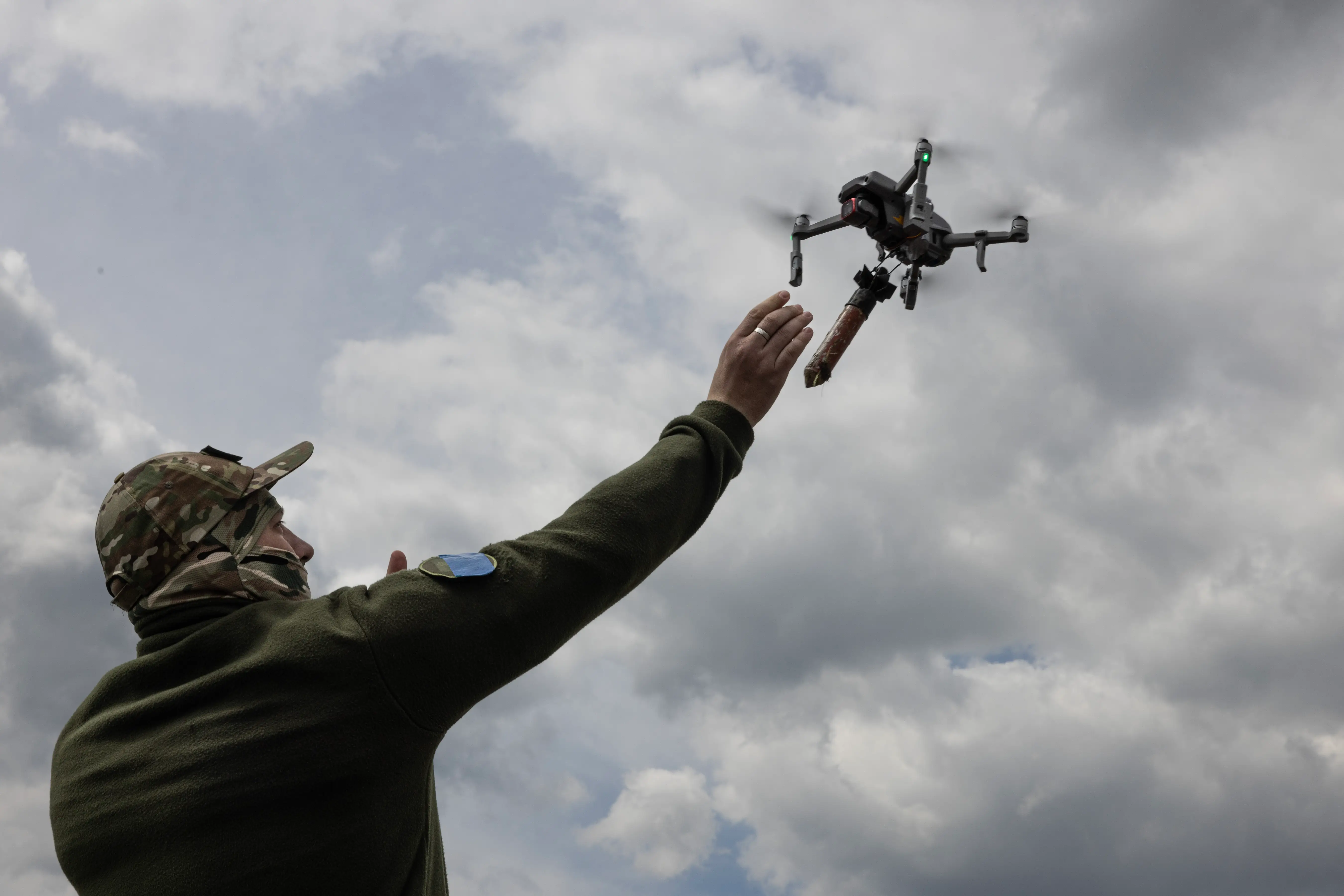 A man in khaki gear extends his arm up to a small drone flying in a blue but cloudy sky
