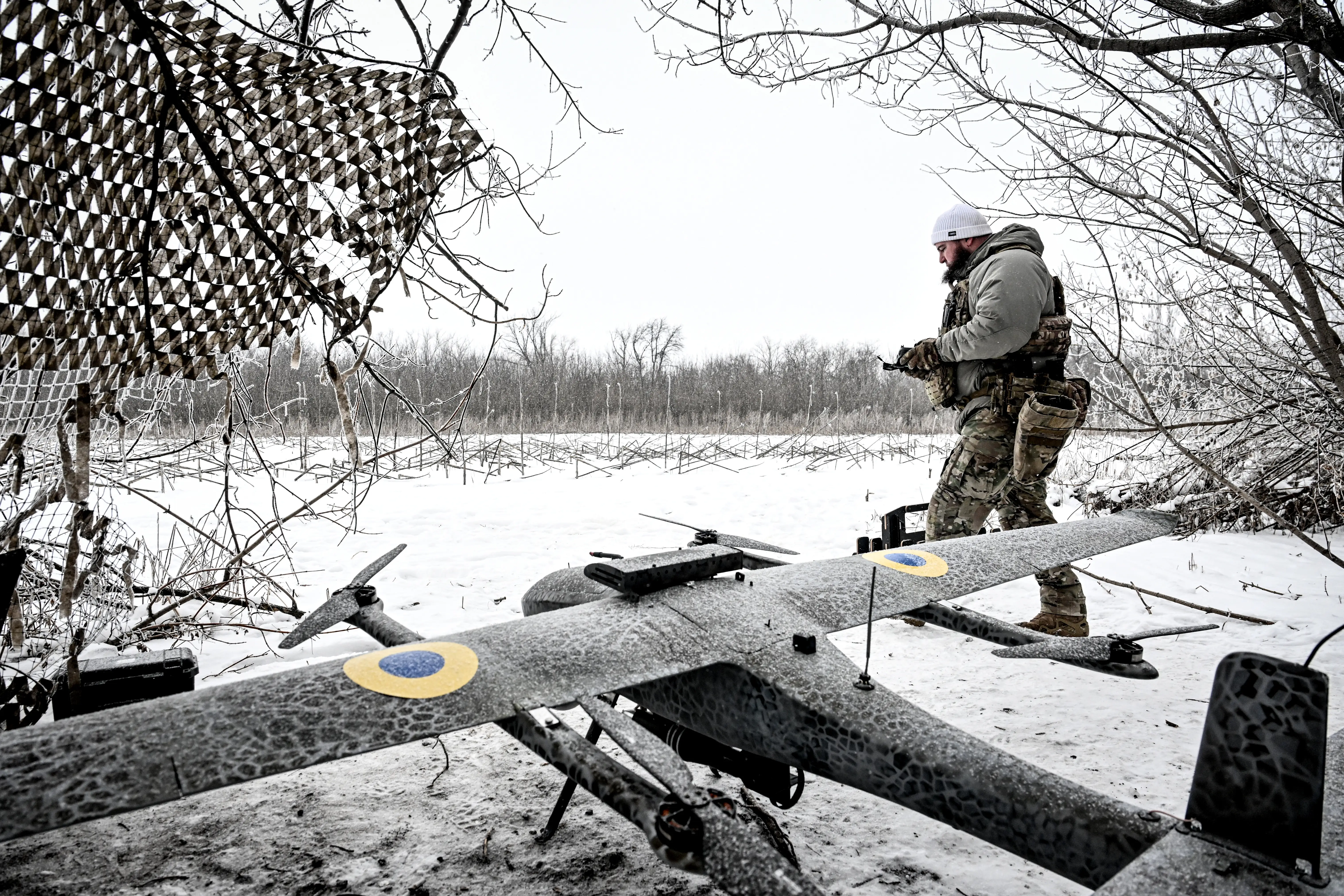 A large grey drone on the ground and covered in snow in a netted position beside a snowy ground and white sky and a soldier in camouflage gear.