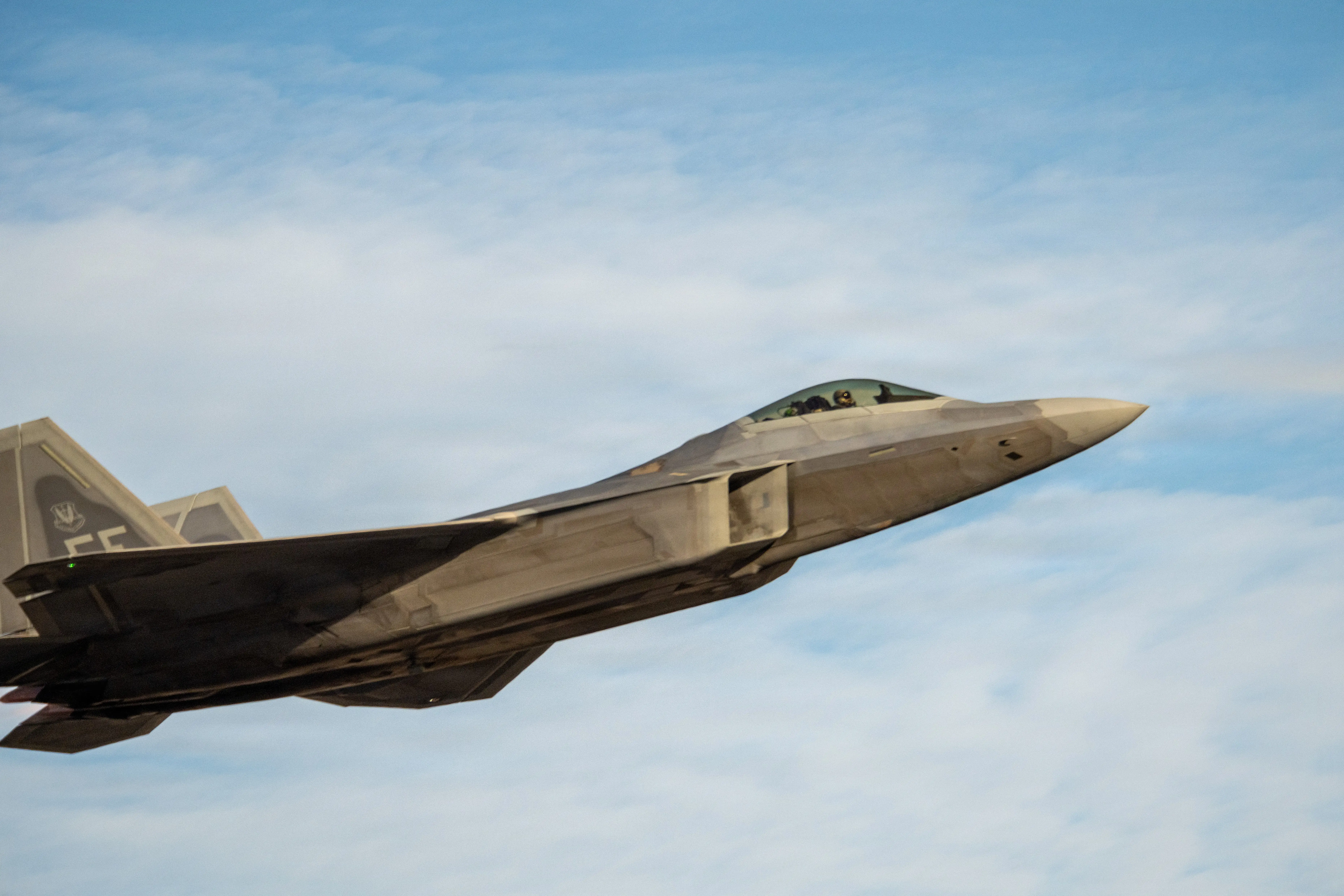 An F-22 flies in a blue and cloudy sky.