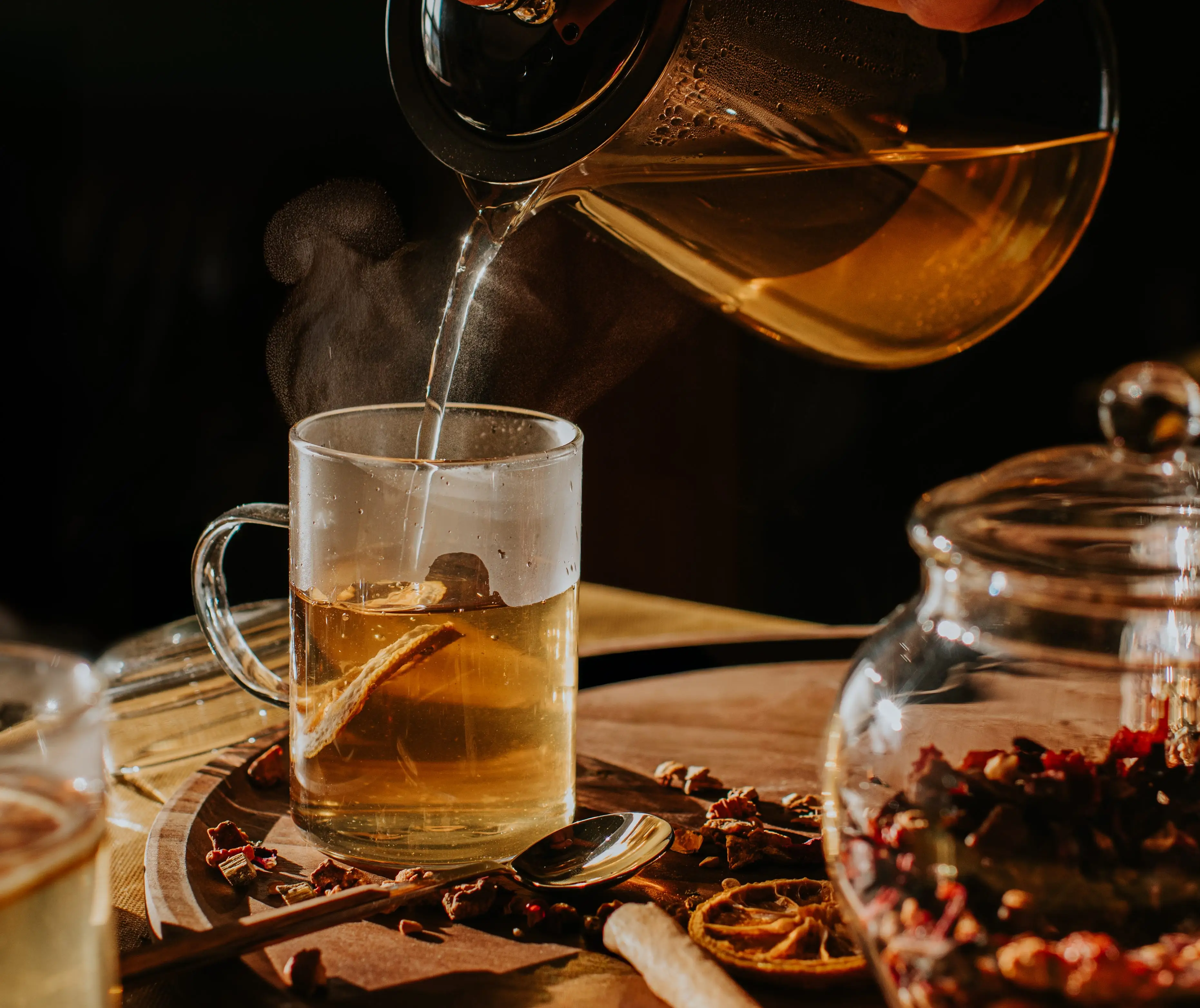 A close up of a cup of herbal tea being poured from a teapot on a tray of dried citrus and spices