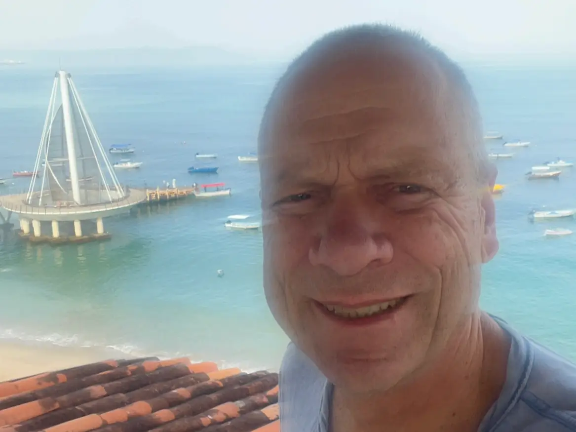Smiling at the camera, a man stands on a balcony overlooking a beach, turquoise water, anchored boats, and a white sail-shaped pier.