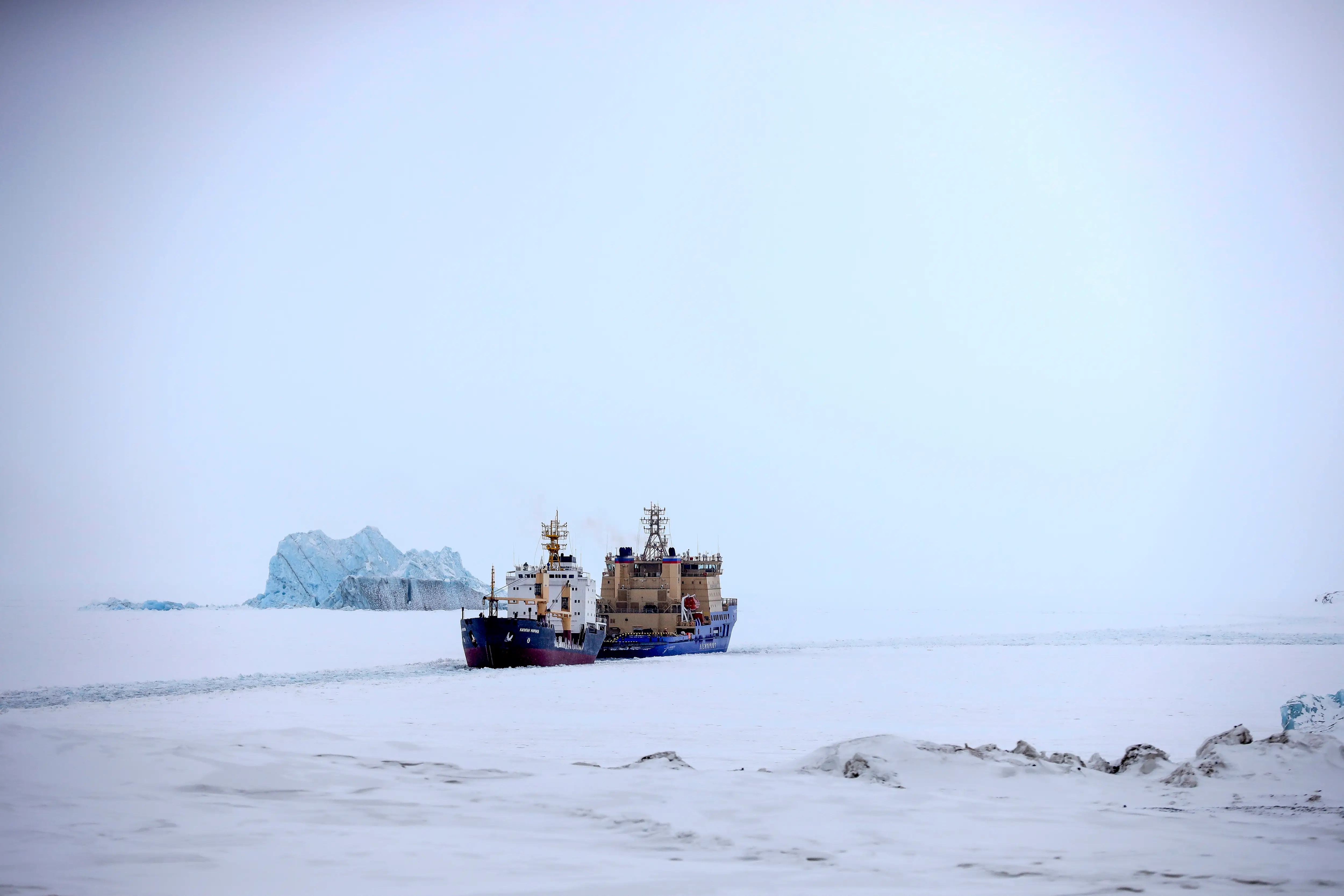 An Icebreaker making the path for a cargo ship with an iceberg in the background near a port on the Alexandra Land island near Nagurskoye, Russia, Monday, May 17, 2021.