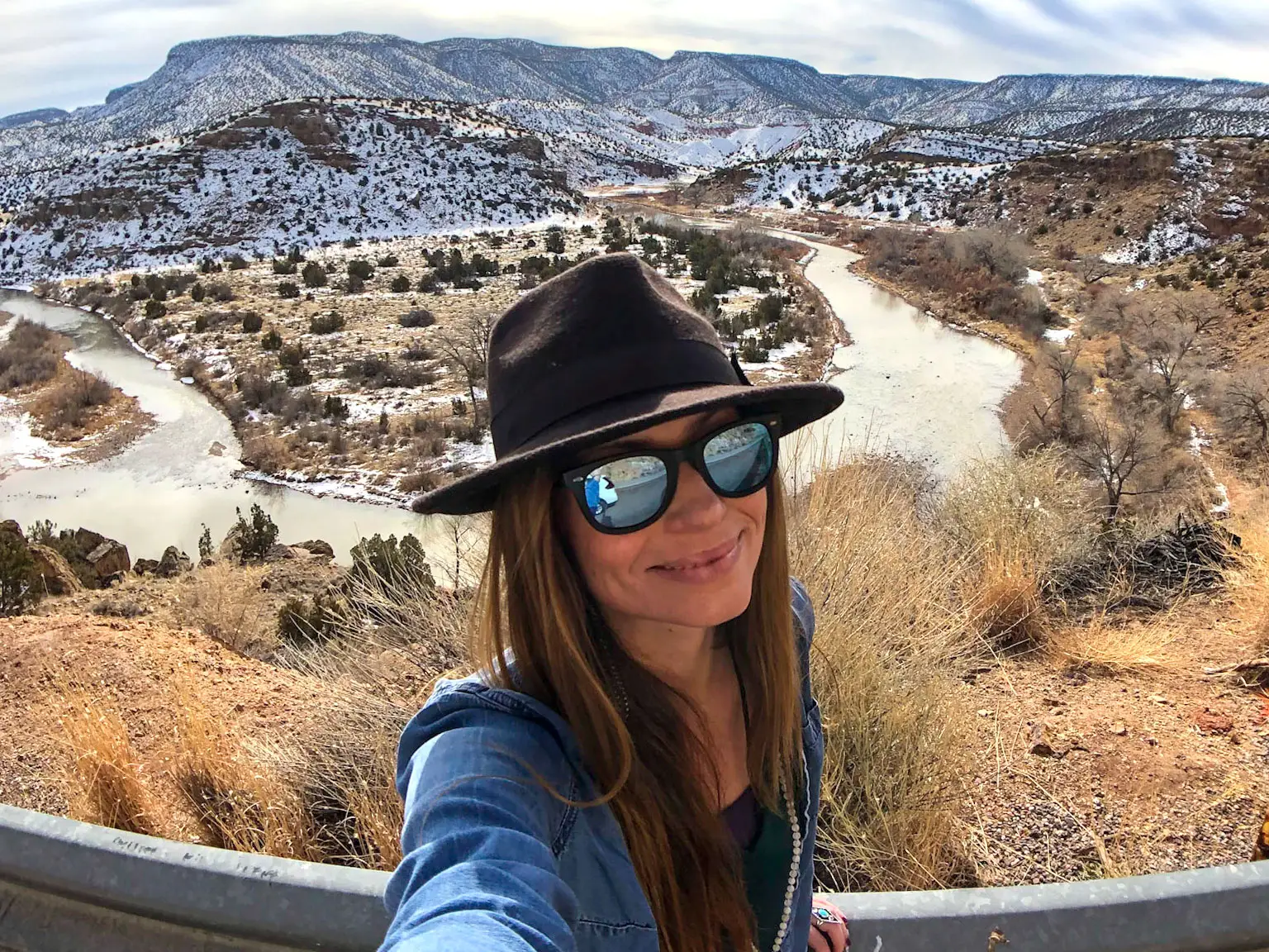 Emily takes a selfie with snow-covered mountains and a river in the background.