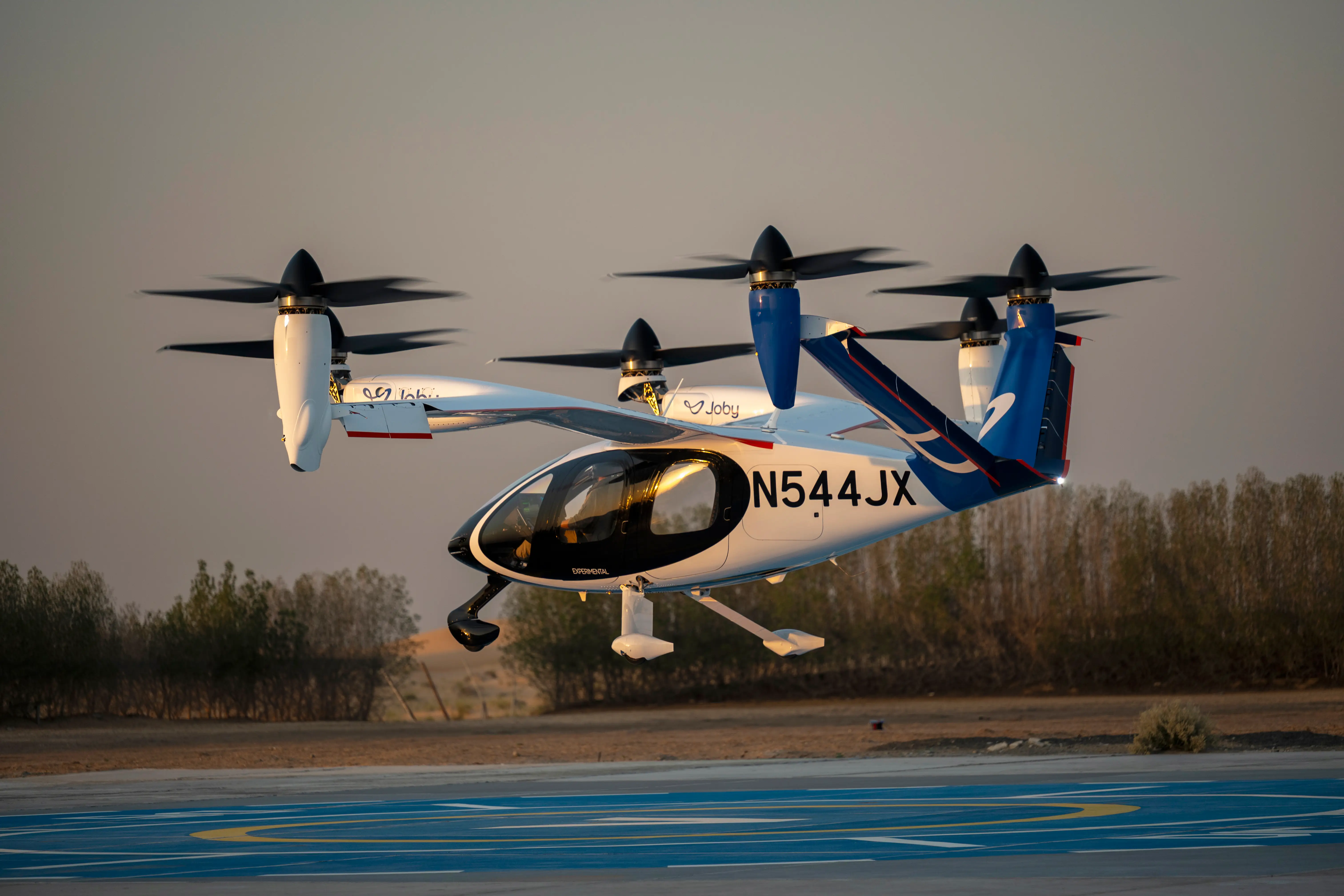 A Joby air taxi hovers above a landing pad, powered by six propellers and with three wheels underneath it.