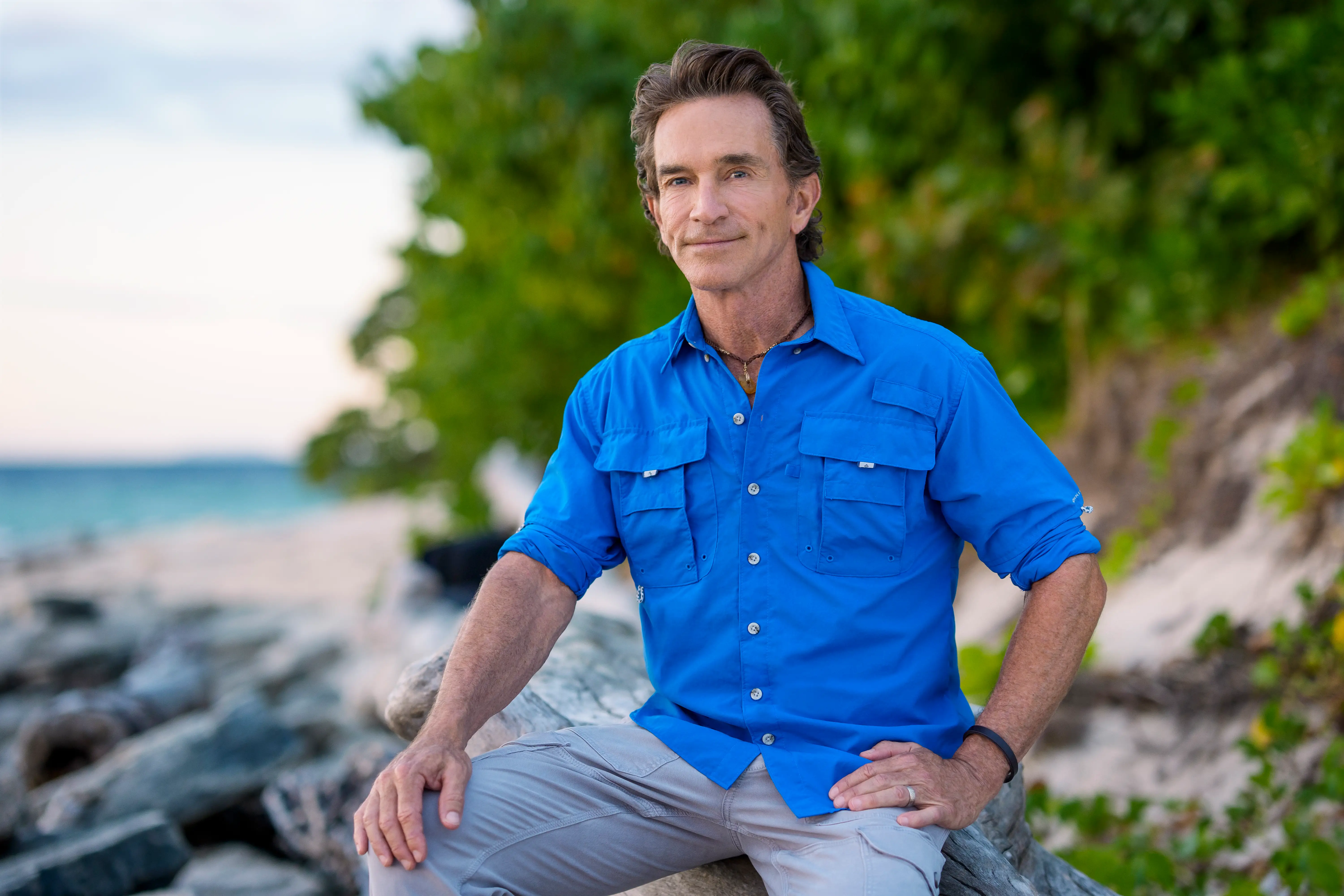 A man sits on a rock on a beach in a blue shirt.