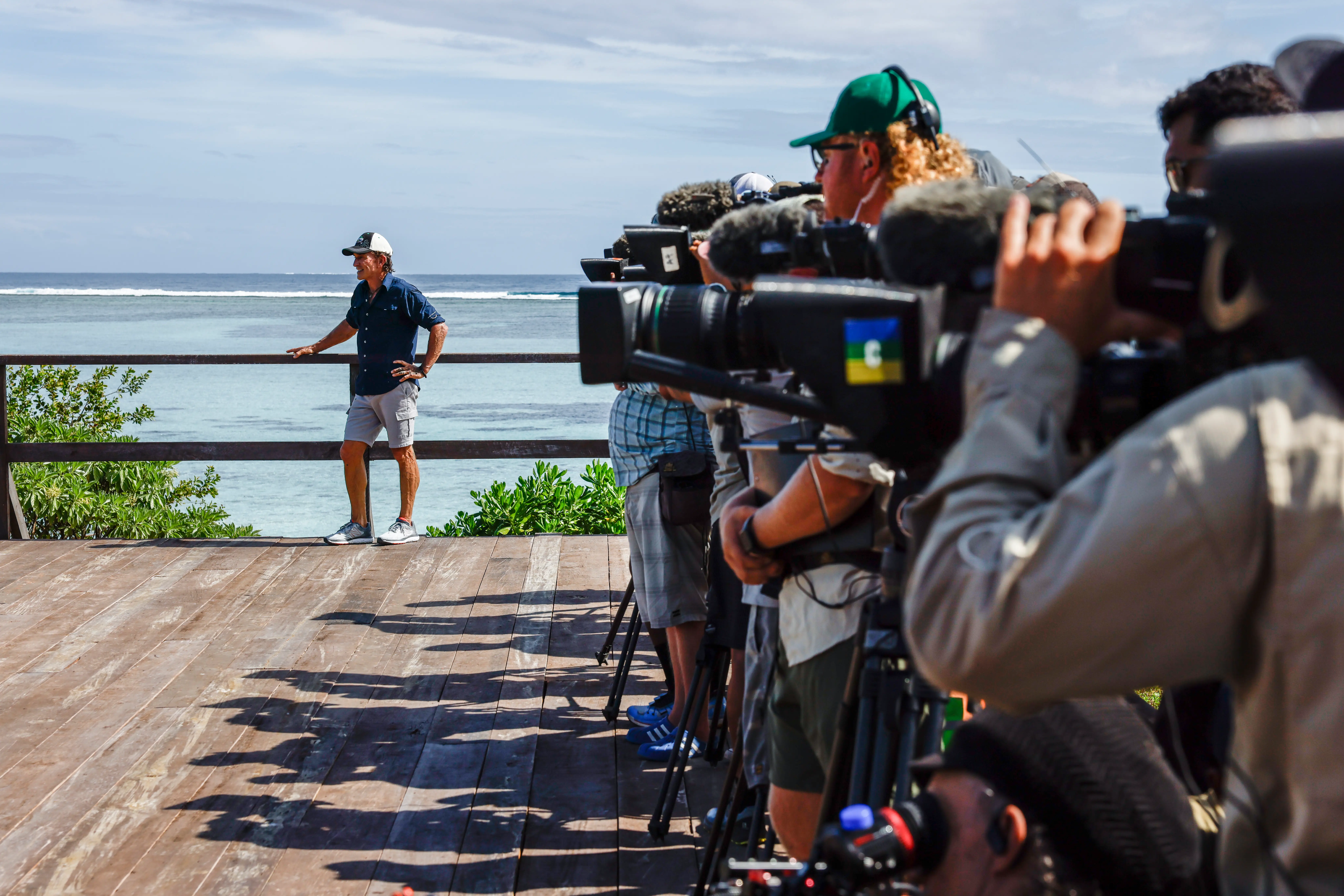 A man stands on a deck by the water, flanked by numerous cameras filming him.
