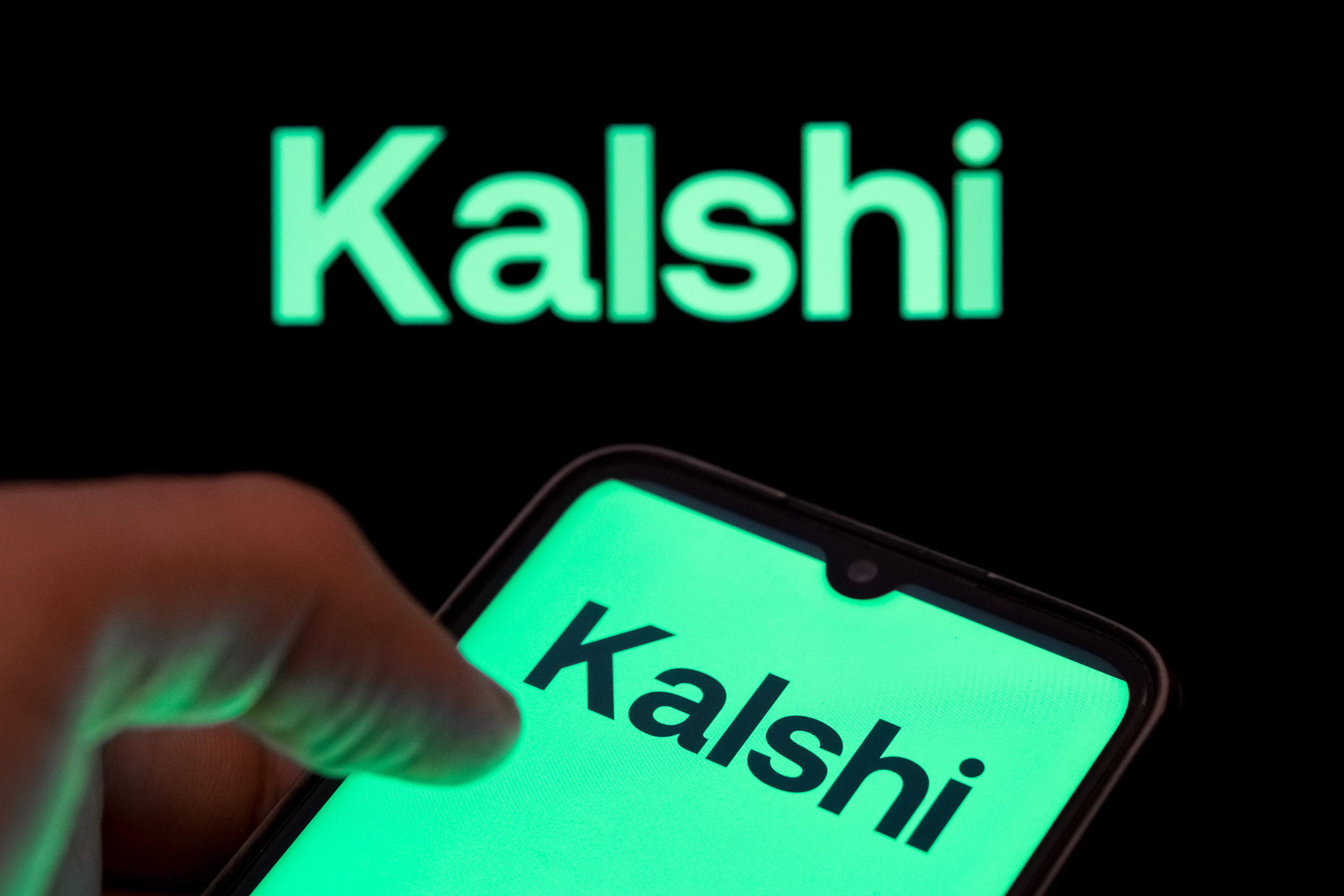 A photo of a person holding a phone with Kalshi's logo on it, with the word