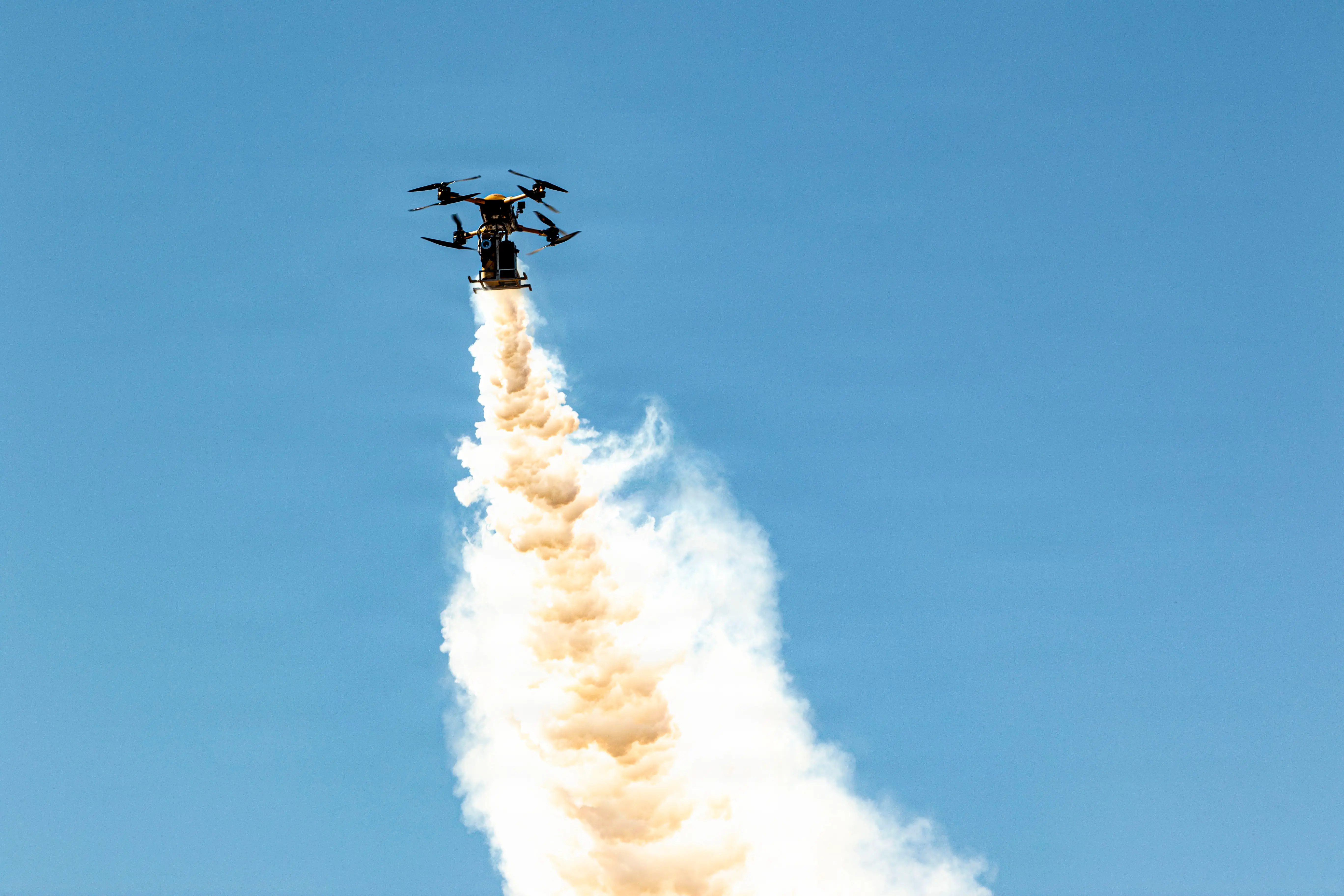 A small quadcopter drone flies in a blue sky with a trail of smoke behind it.