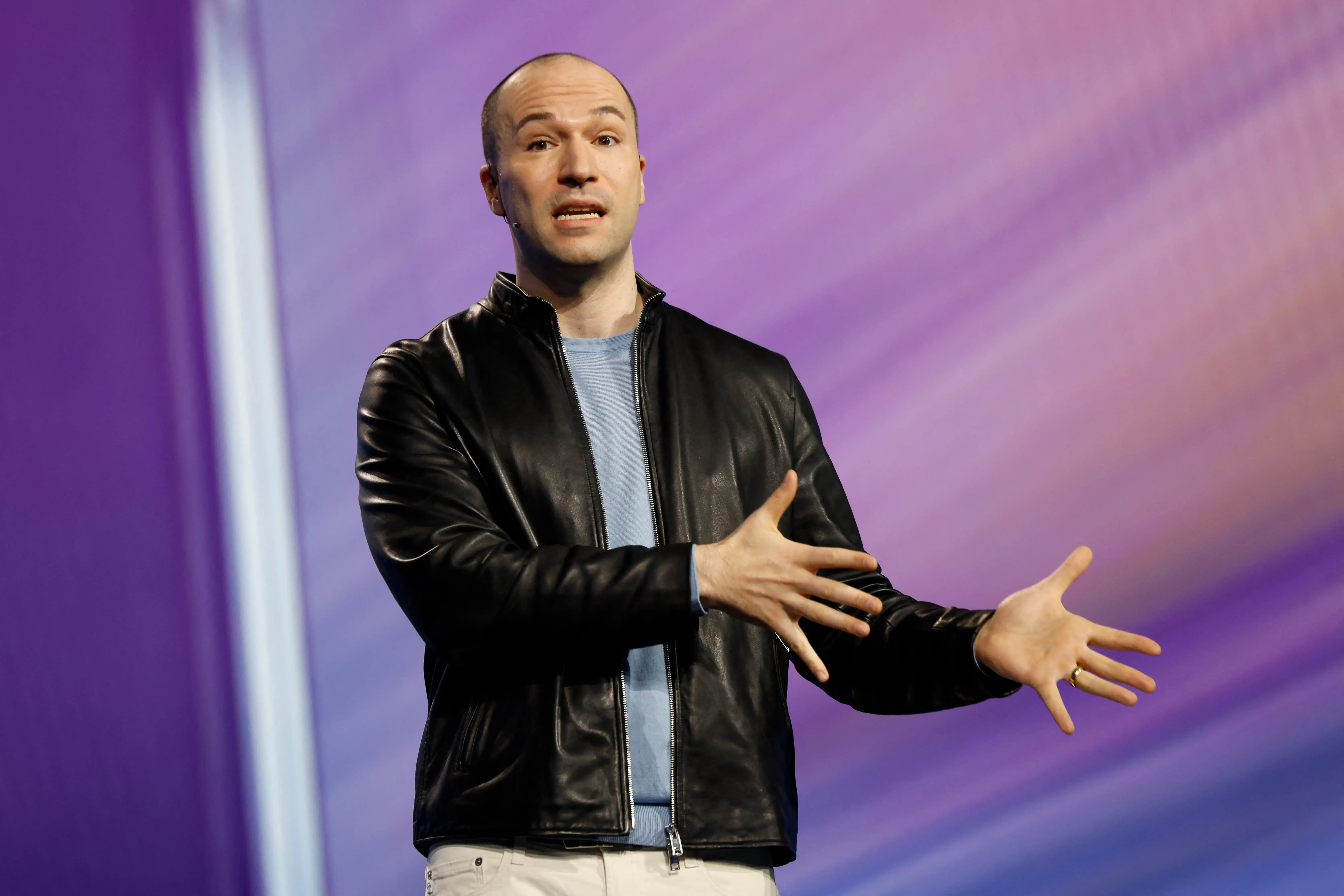 Greg Brockman, wearing a black leather jacket and a blue shirt, is standing on stage giving a speech. His hands are both splayed to his left.
