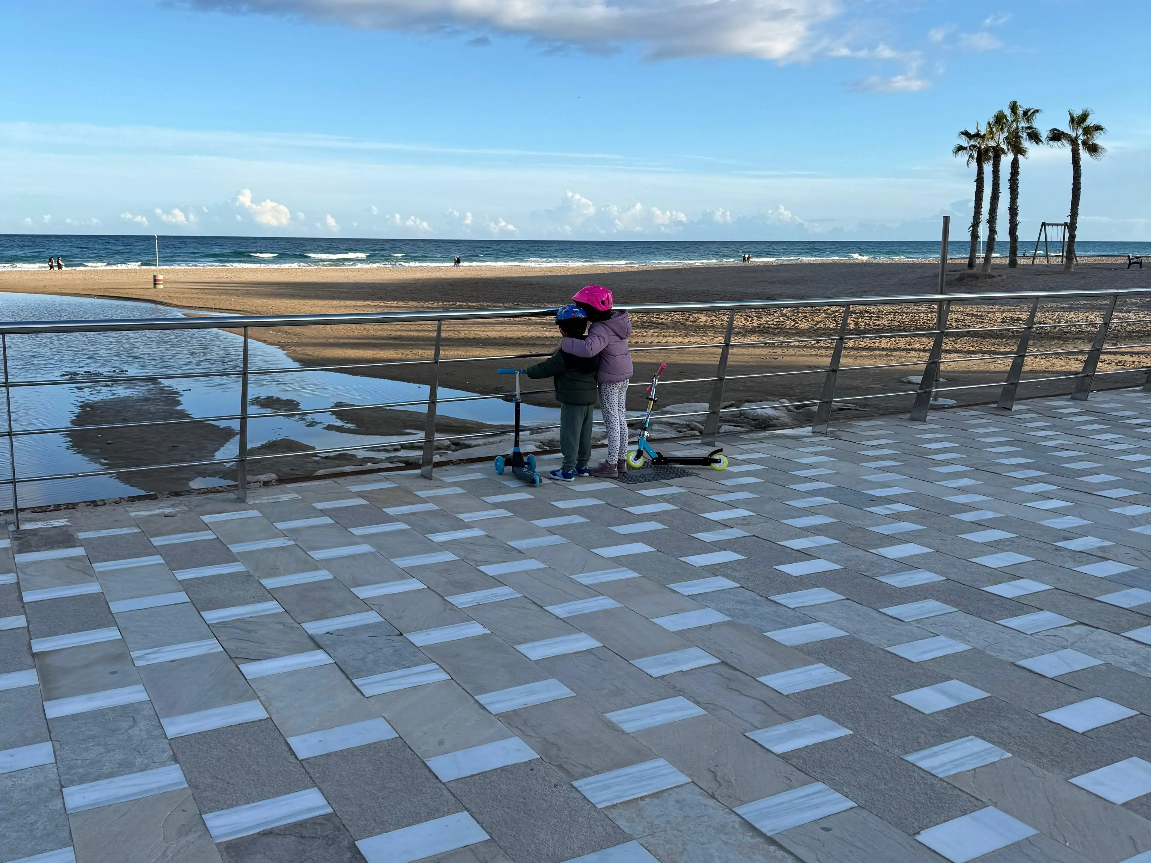 Two children standing next to scooters look out at the ocean.