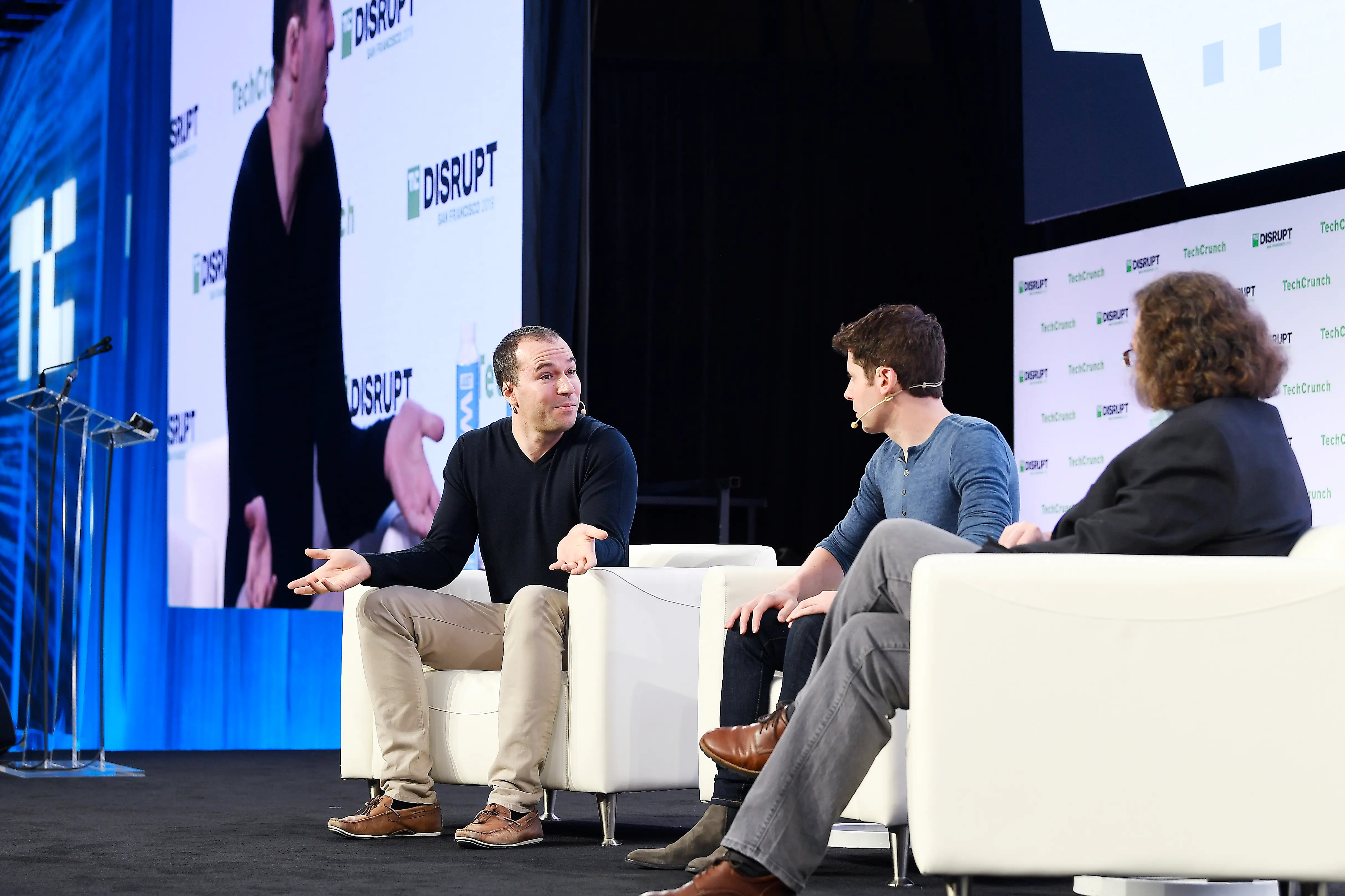 Sam Altman is sitting on a white chair on a stage during a TechCrunch event. He is next to Sam Altman.