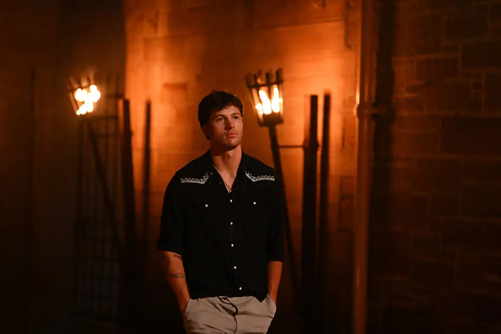 A man in a black short sleeved Western shirt and brown hair stands in front of a stone wall with two torches behind him.