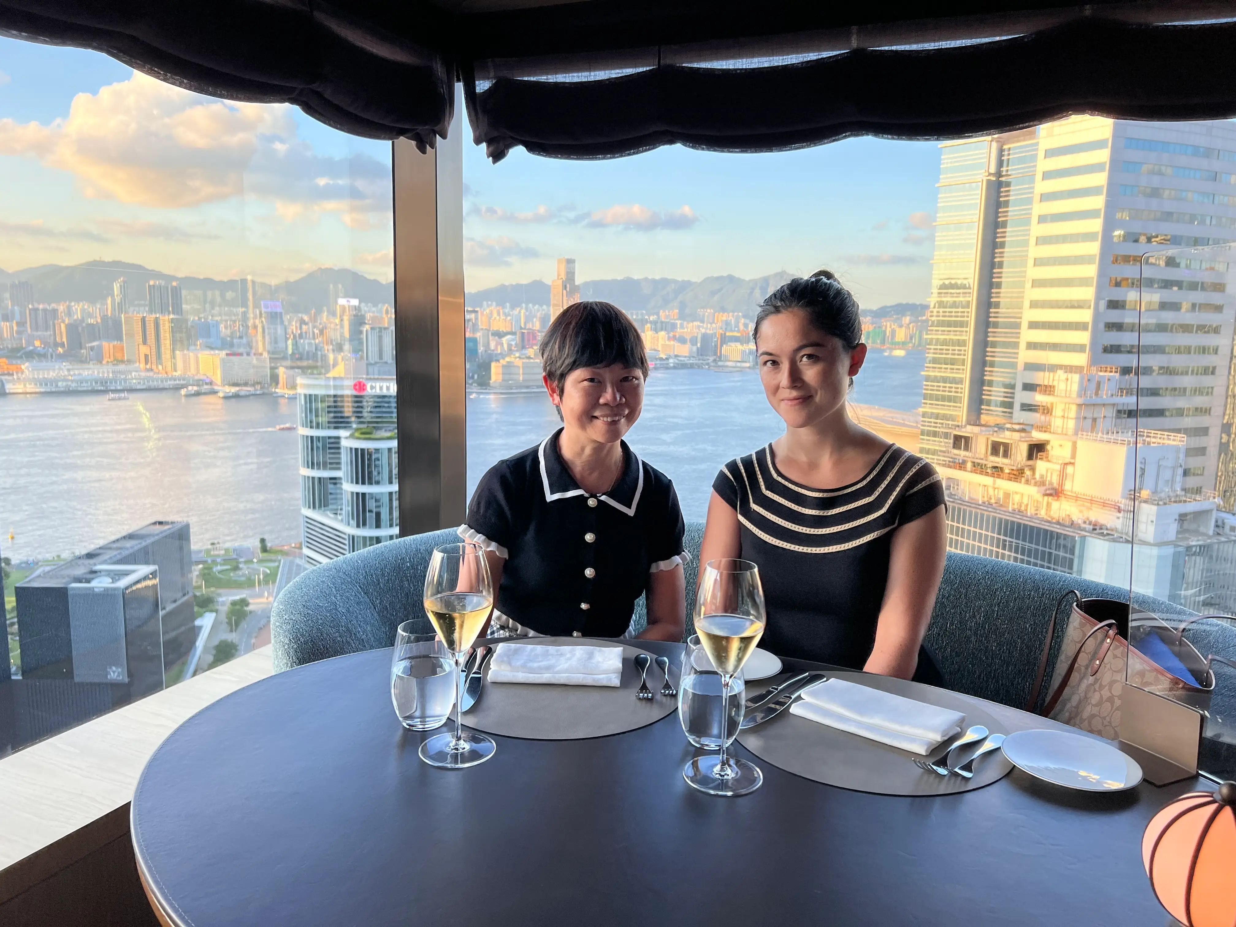 Mother and daughter posing with two glasses of white wine and Hong Kong skyline behind them.