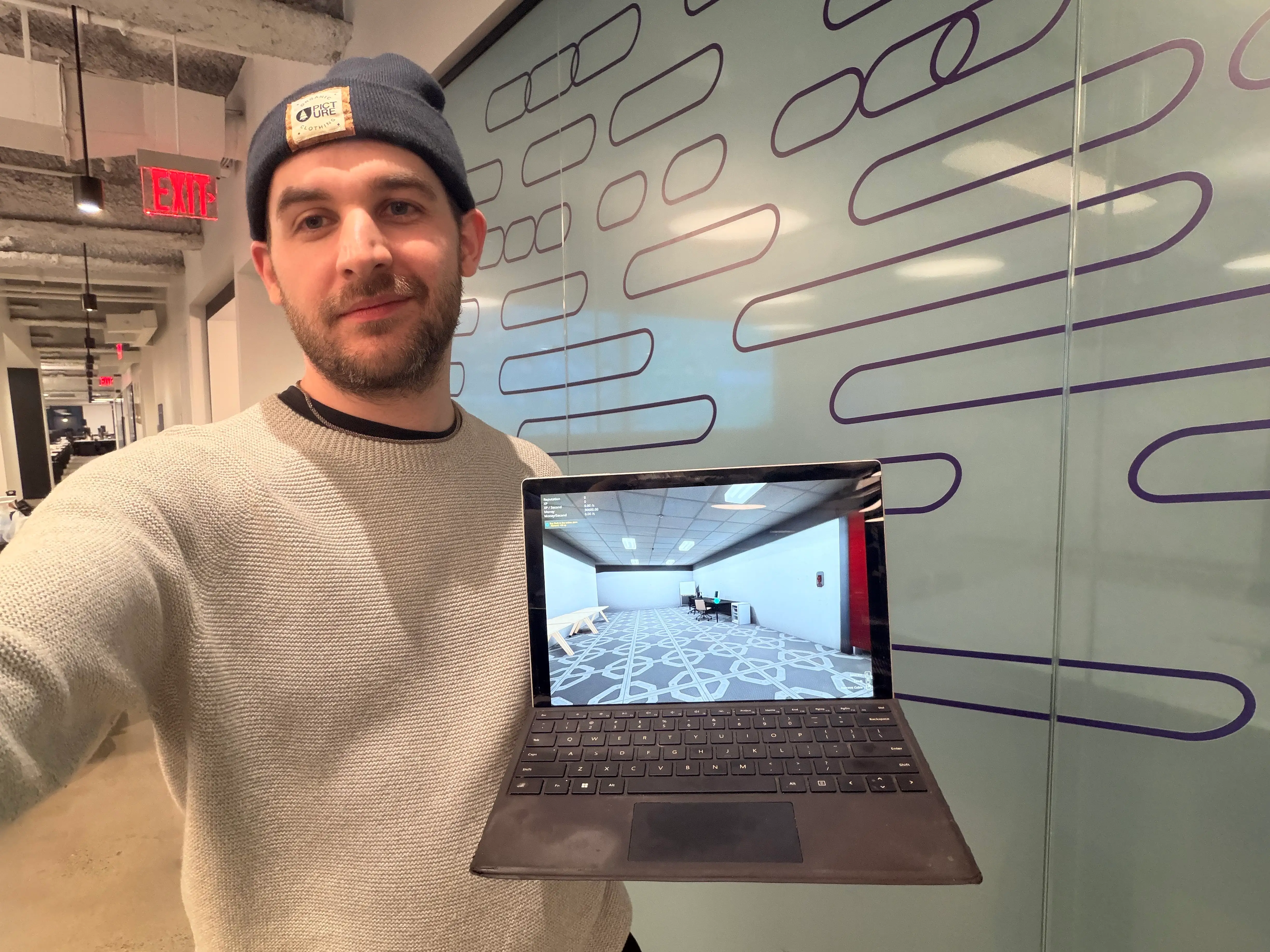 A picture of journalist Ben Shimkus in a grey sweater and blue hat. He's holding a computer that's playing the 'Data Center' demo game on Stream.