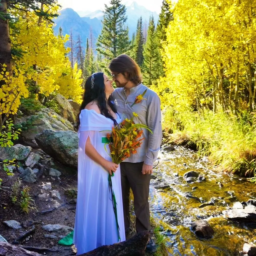 A couple stands by a stream in an autumn forest with bright yellow leaves, looking into each other's eyes.